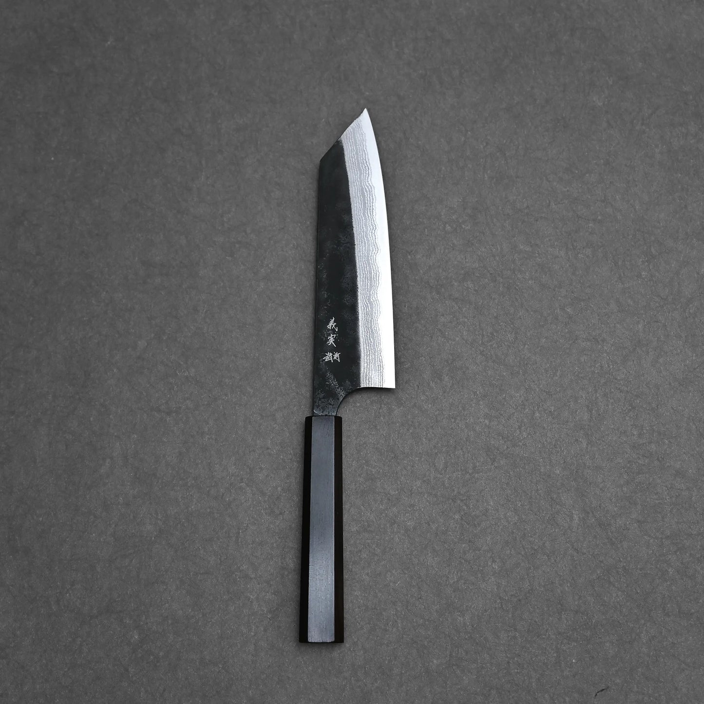 A Yoshimi Kato kurouchi damascus AS bunka 170mm knife with a straight-edged, patterned Aogami Super steel blade and Japanese characters by the black handle rests on a dark grey textured surface.