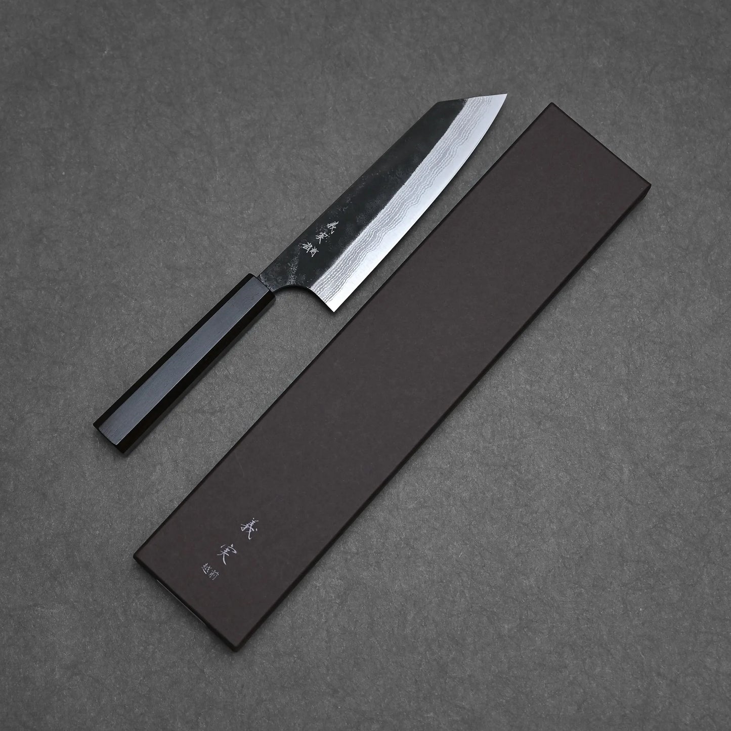 The Yoshimi Kato kurouchi damascus AS bunka 170mm, featuring a black handle and patterned blade, rests beside a dark knife box with white text, all displayed on a textured gray surface.