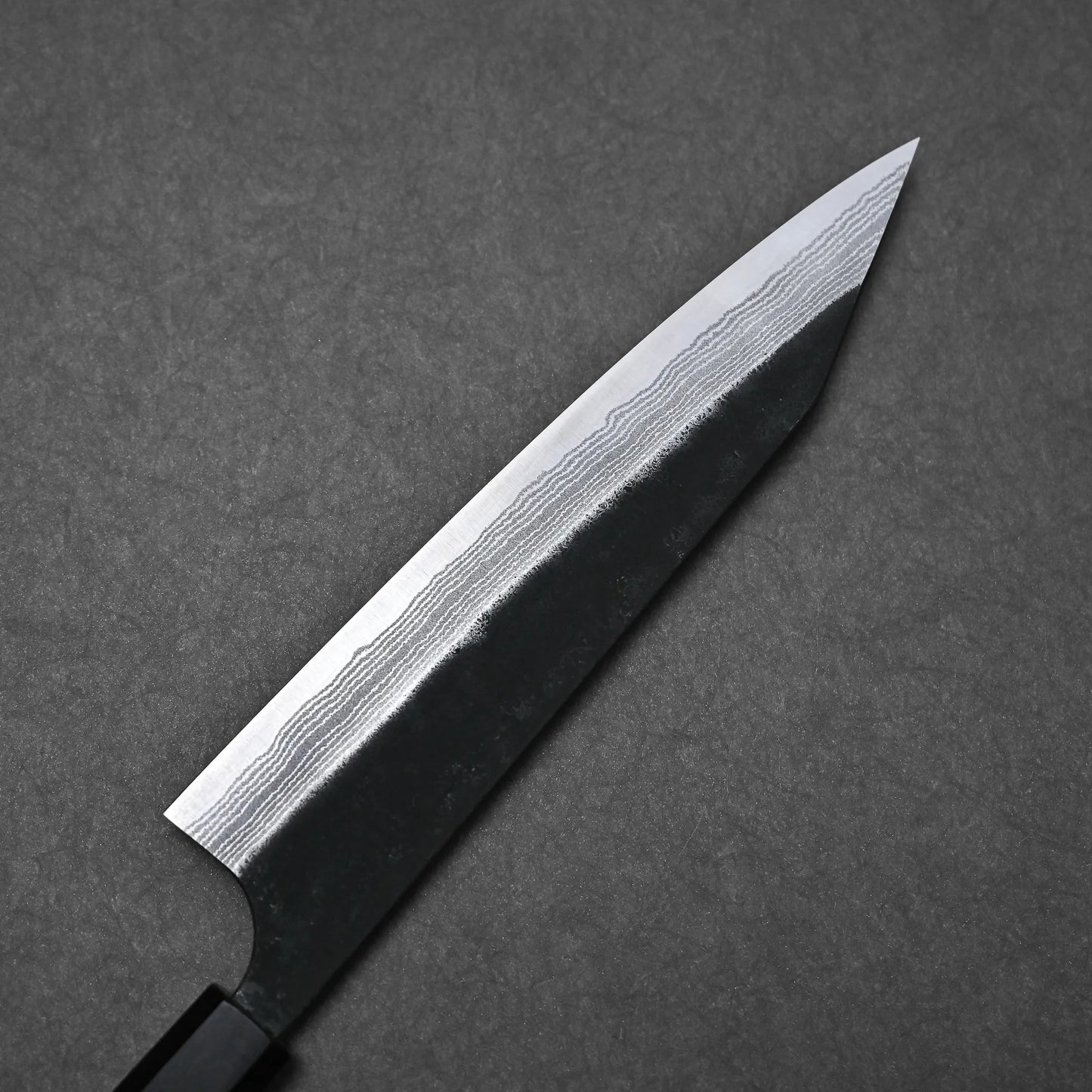 A close-up of the Yoshimi Kato kurouchi damascus AS kiritsuke gyuto 210mm showcases its layered blade pattern, sharp tip, and polished edge set against a dark textured background.