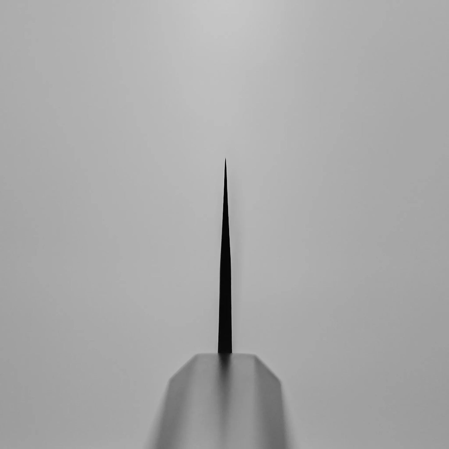 Close-up, symmetrical view of the Yoshimi Kato kurouchi damascus AS kiritsuke gyuto 210mm blade tip, centered and in focus against a plain light grey background; the knife’s edge is sharp while the rest appears blurred.