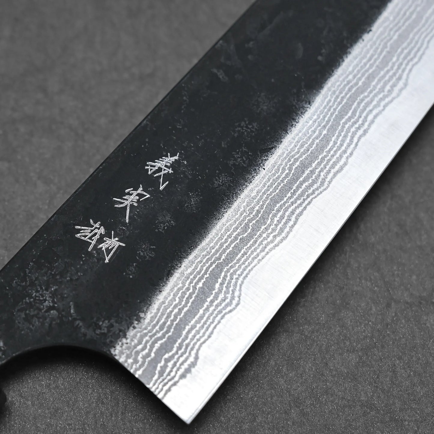 Close-up of a Yoshimi Kato kurouchi damascus AS kiritsuke gyuto 210mm blade, highlighting wavy Damascus layers and kanji engraving on a dark surface.
