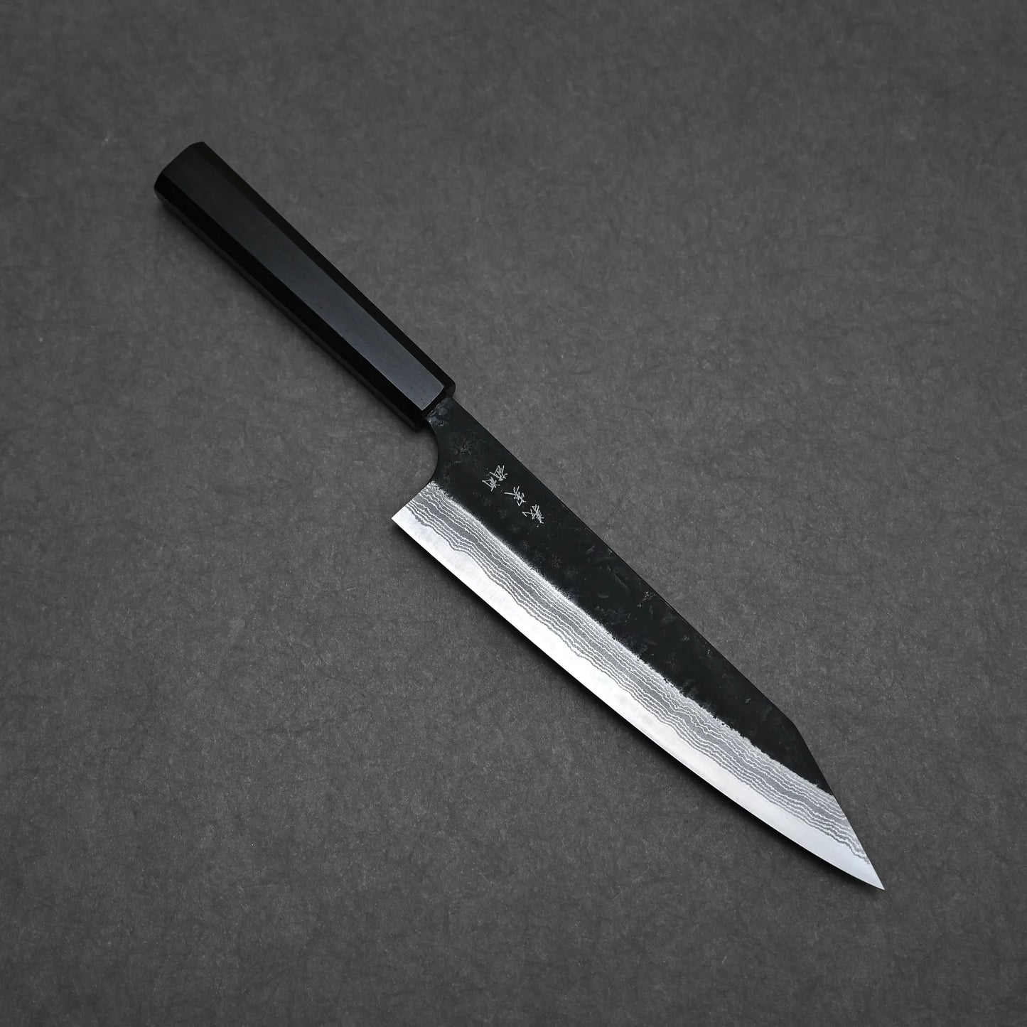 The Yoshimi Kato kurouchi damascus AS kiritsuke gyuto 210mm, with a dark handle and patterned angular blade, is displayed on a dark gray surface, showcasing visible engravings near the spine and refined craftsmanship.