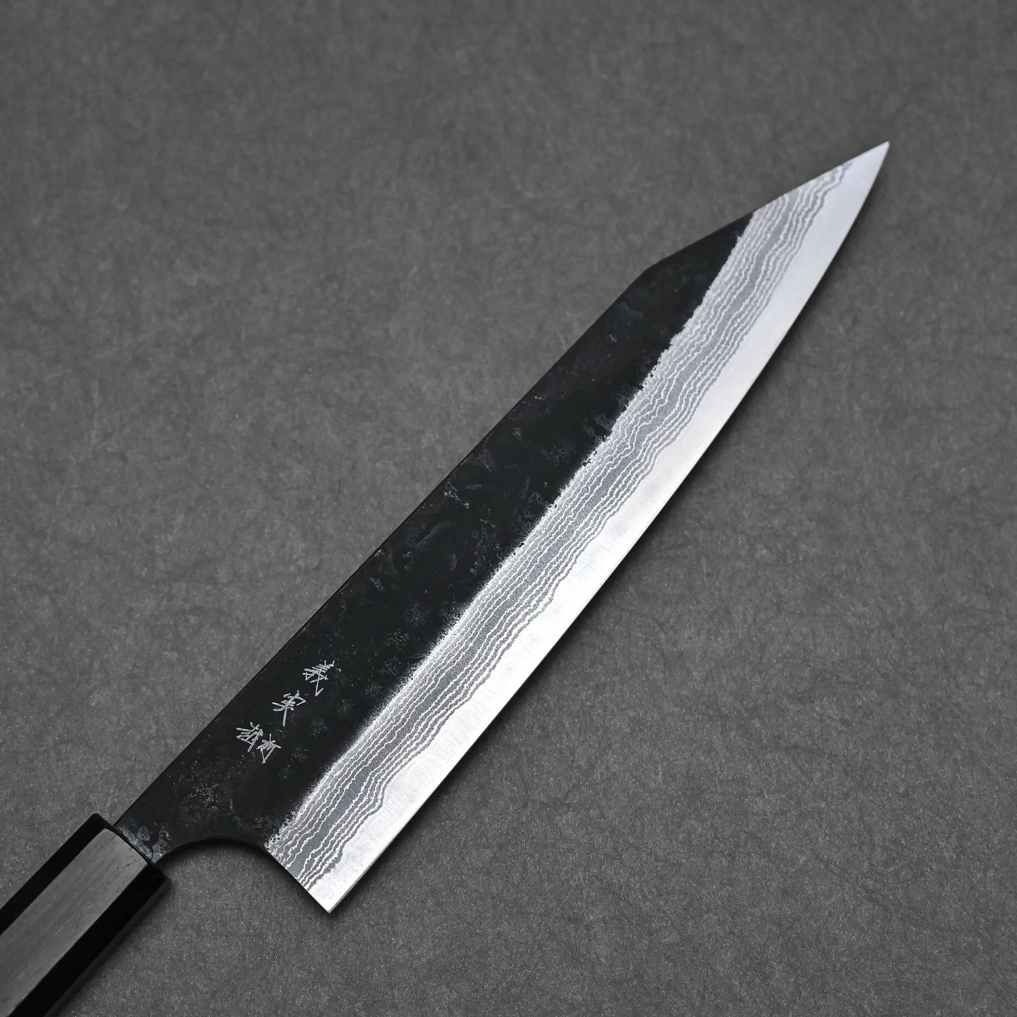The Yoshimi Kato kurouchi damascus AS kiritsuke gyuto 210mm features a dark handle, Aogami Super blade with wavy damascus patterns, and Japanese characters on the metal, displayed on a textured gray surface.