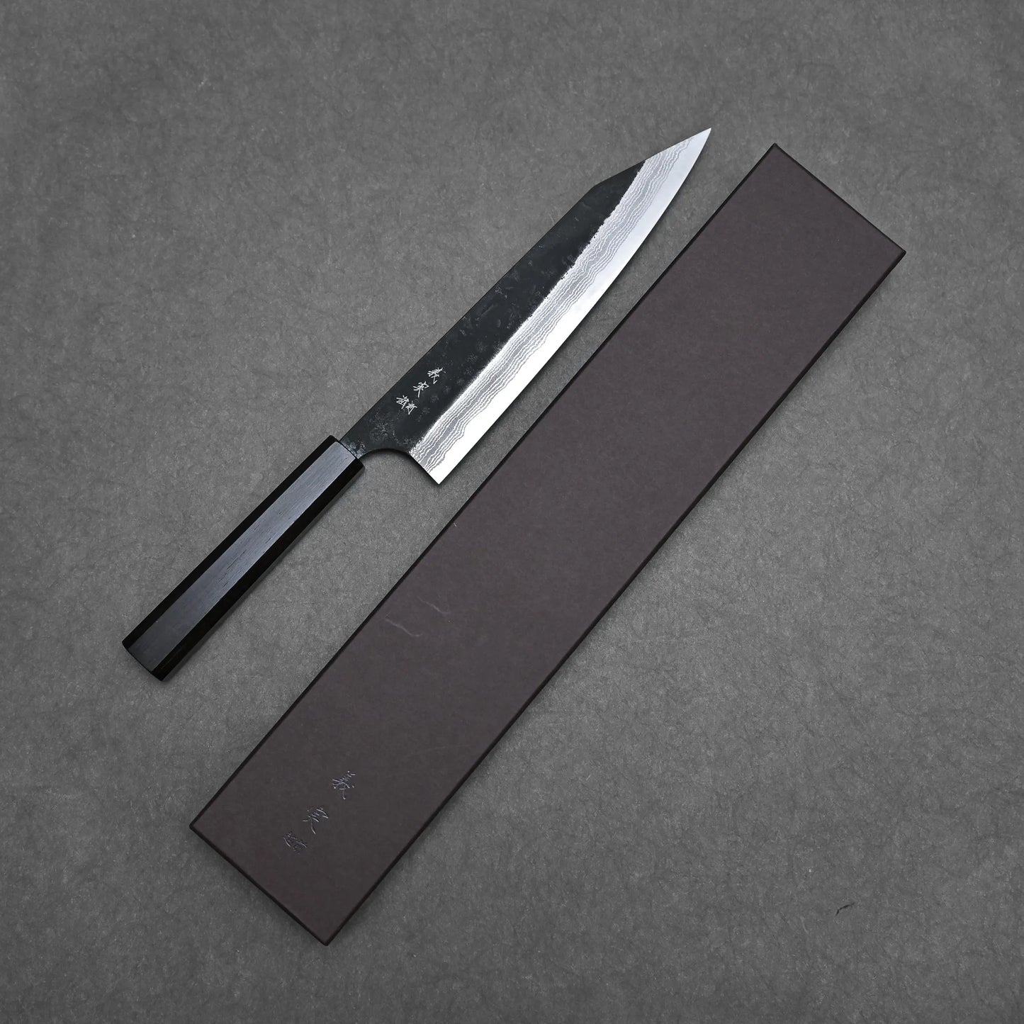 The Yoshimi Kato kurouchi damascus AS kiritsuke gyuto 210mm, featuring a dark handle and sharp pointed blade, is placed above its long rectangular case on a gray textured background.