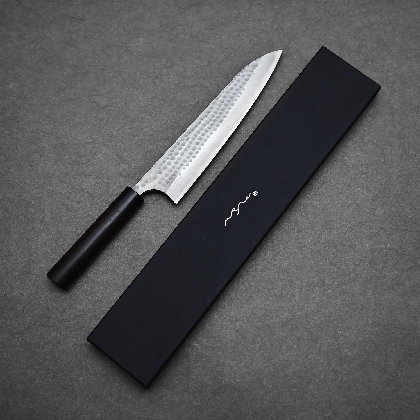 An Anryu tsuchime aogami#2 gyuto 210mm chef knife by Anryu, featuring a hammered carbon steel blade and dark handle, rests beside a sleek black box with minimalist white markings on a textured gray surface.