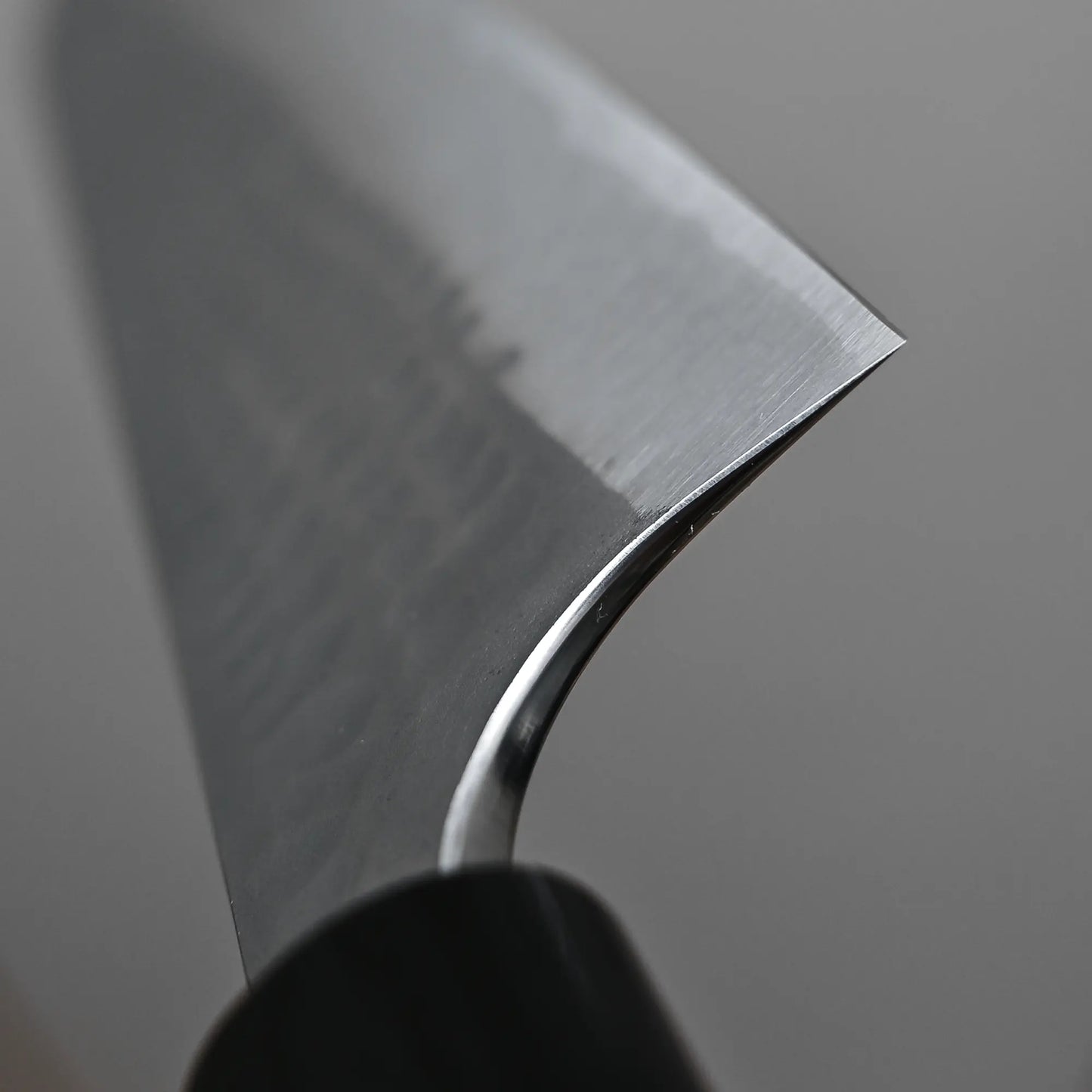 Close-up of the Anryu tsuchime aogami#2 gyuto 210mm by Anryu, featuring a sharp Aogami #2 carbon steel blade with polished, textured edge and reflective finish against a blurred gray background.
