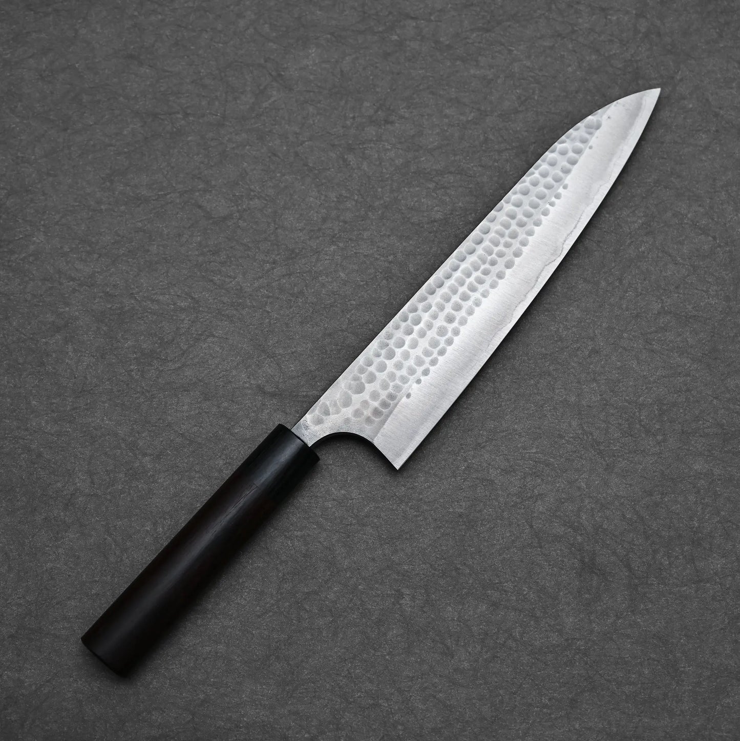 An Anryu tsuchime aogami#2 gyuto 210mm chef knife by Anryu, with a hammered Aogami #2 carbon steel blade and dark wooden handle, is displayed diagonally on a gray textured surface.