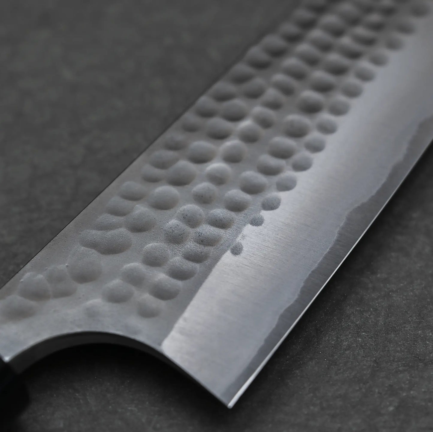 Close-up of the Anryu tsuchime aogami#2 gyuto 210mm by Anryu, featuring a hammered texture on the blade’s upper part and a sharp Aogami #2 carbon steel edge, set on a dark, textured surface.