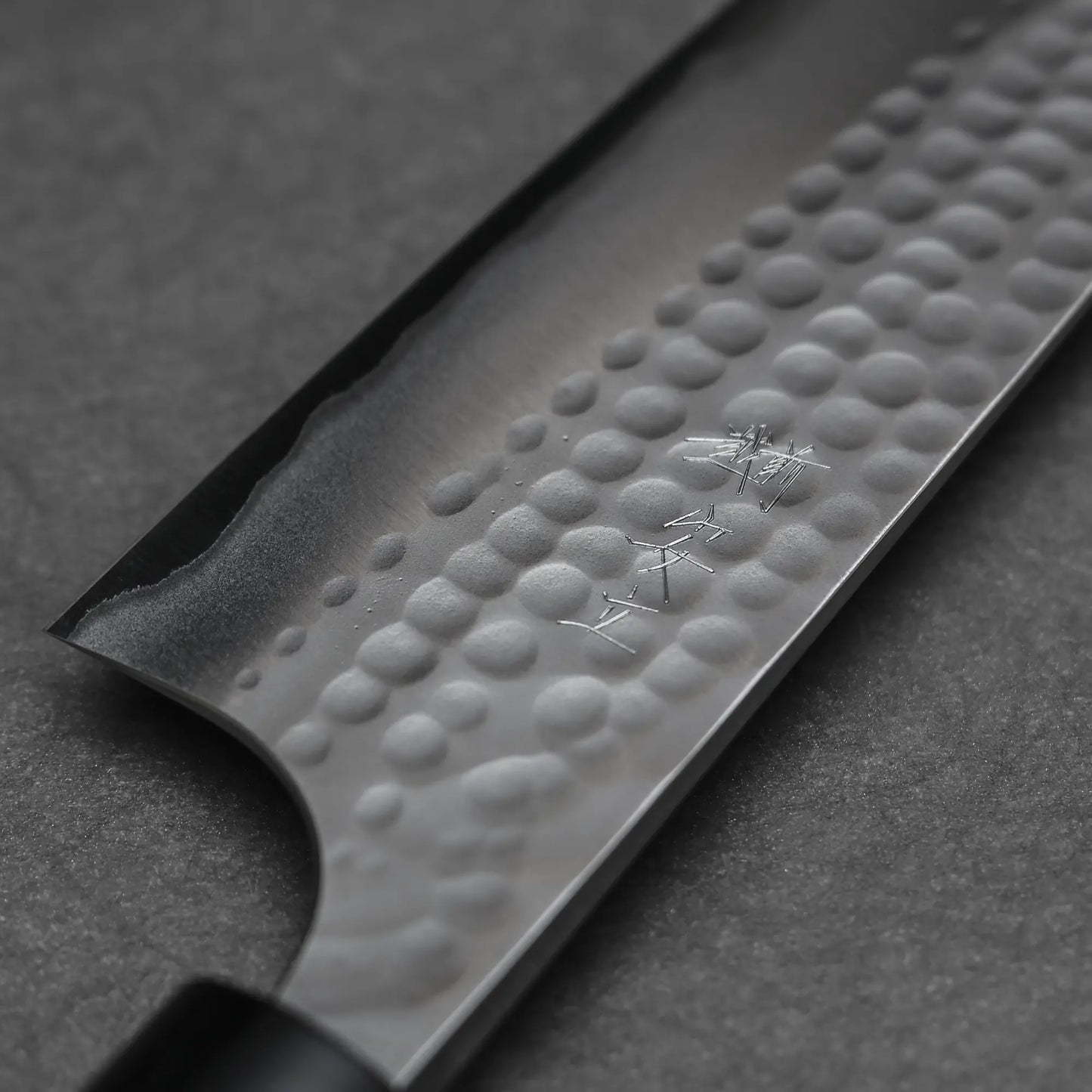 Close-up of the Anryu tsuchime aogami#2 gyuto 210mm by Anryu, showcasing its hammered Aogami #2 carbon steel blade with kanji engravings, set against a dark, textured background.