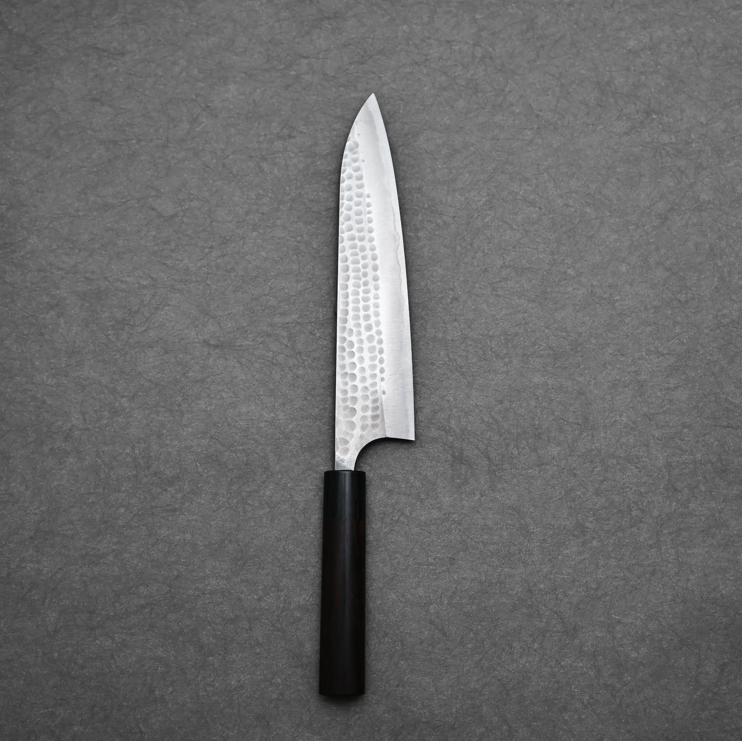 The Anryu tsuchime aogami#2 gyuto 210mm by Anryu, featuring a hammered Aogami #2 carbon steel blade and dark wooden handle, is placed on a textured gray surface.