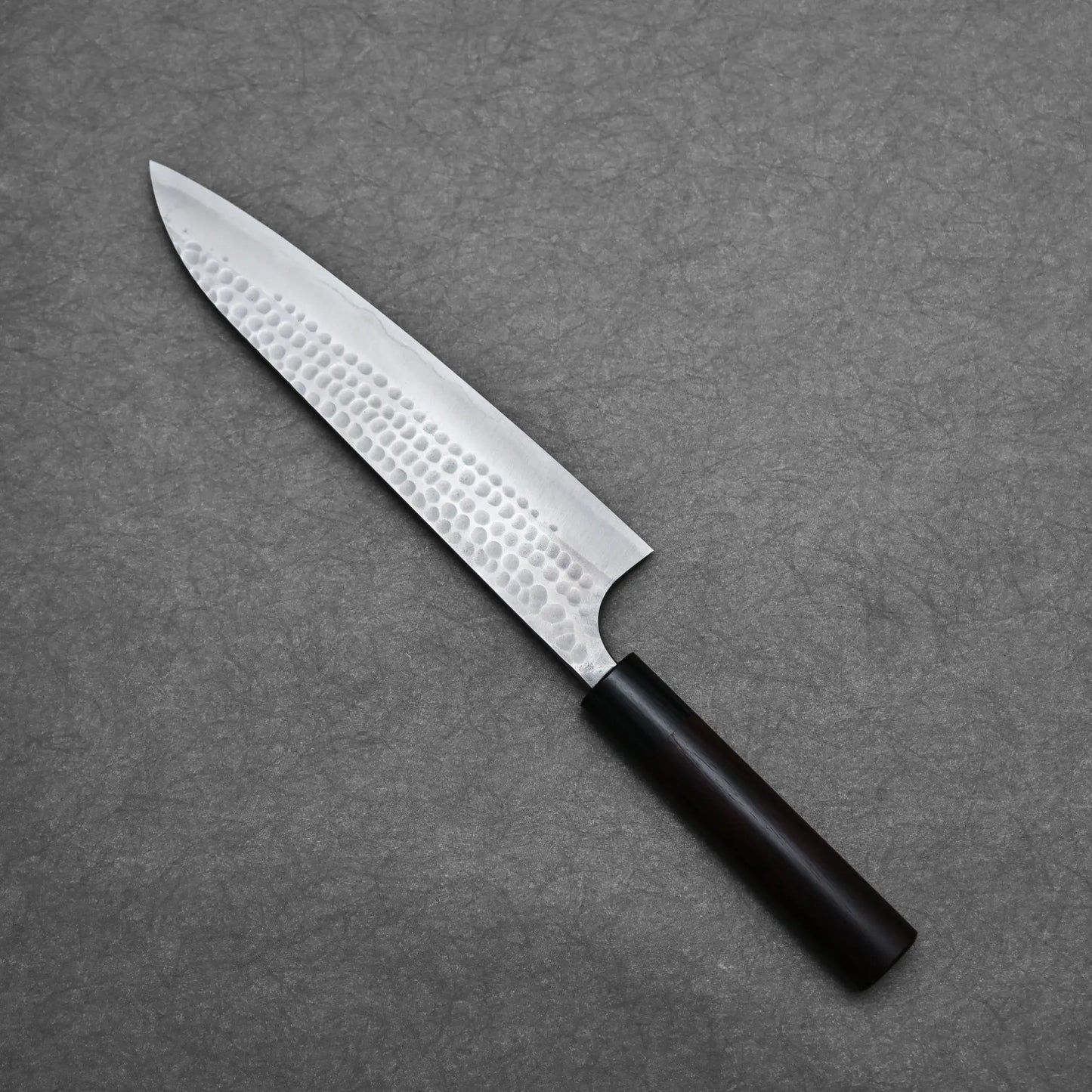 The Anryu tsuchime aogami#2 gyuto 210mm by Anryu features a hammered Aogami #2 carbon steel blade and a dark wooden handle, beautifully displayed on a gray textured surface.