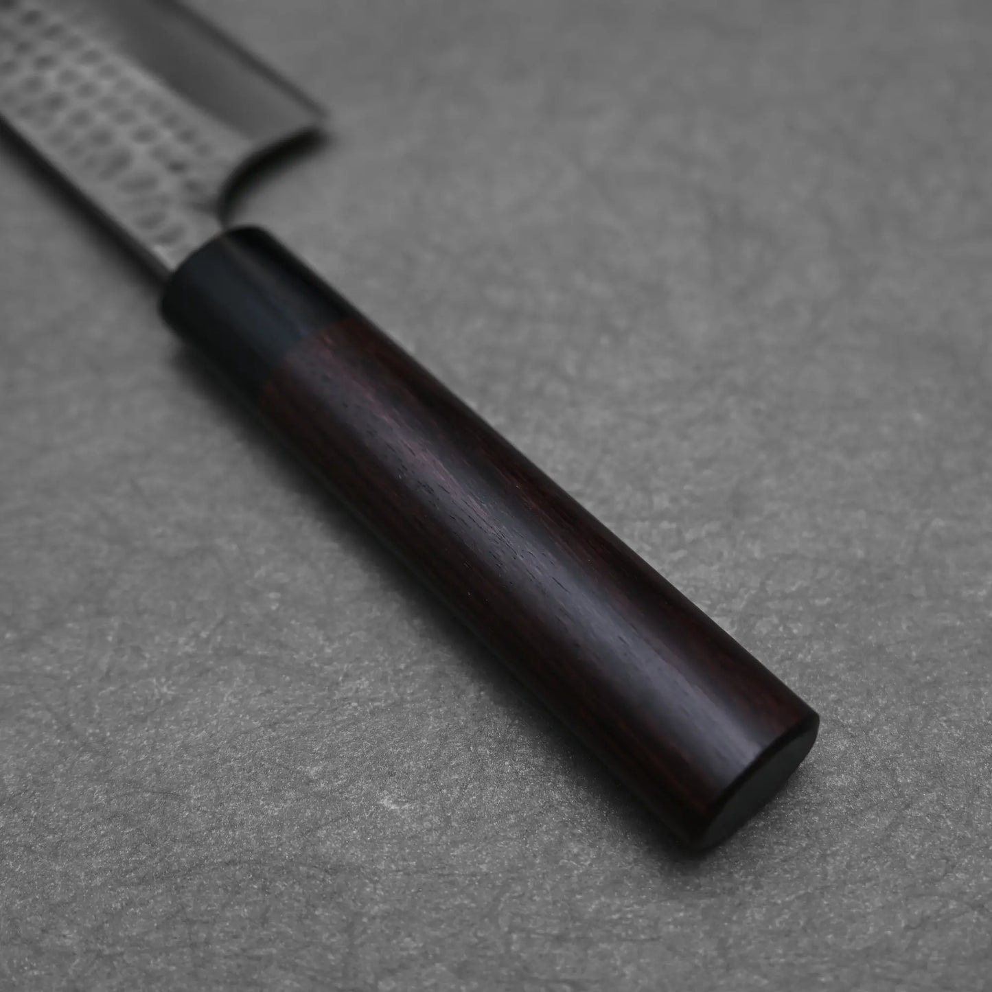 Close-up of an Anryu tsuchime aogami#2 gyuto 210mm chef knife by Anryu, showing its dark wood handle and part of the Aogami #2 carbon steel blade with textured bolster, resting on a gray textured surface.