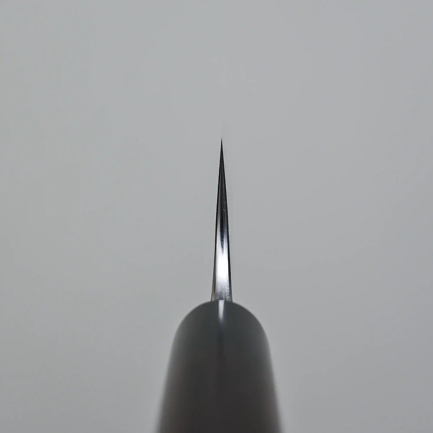 Close-up of the sharp tip of the Anryu tsuchime aogami#2 gyuto 210mm by Anryu, crafted from Aogami #2 carbon steel, positioned vertically against a plain gray background.