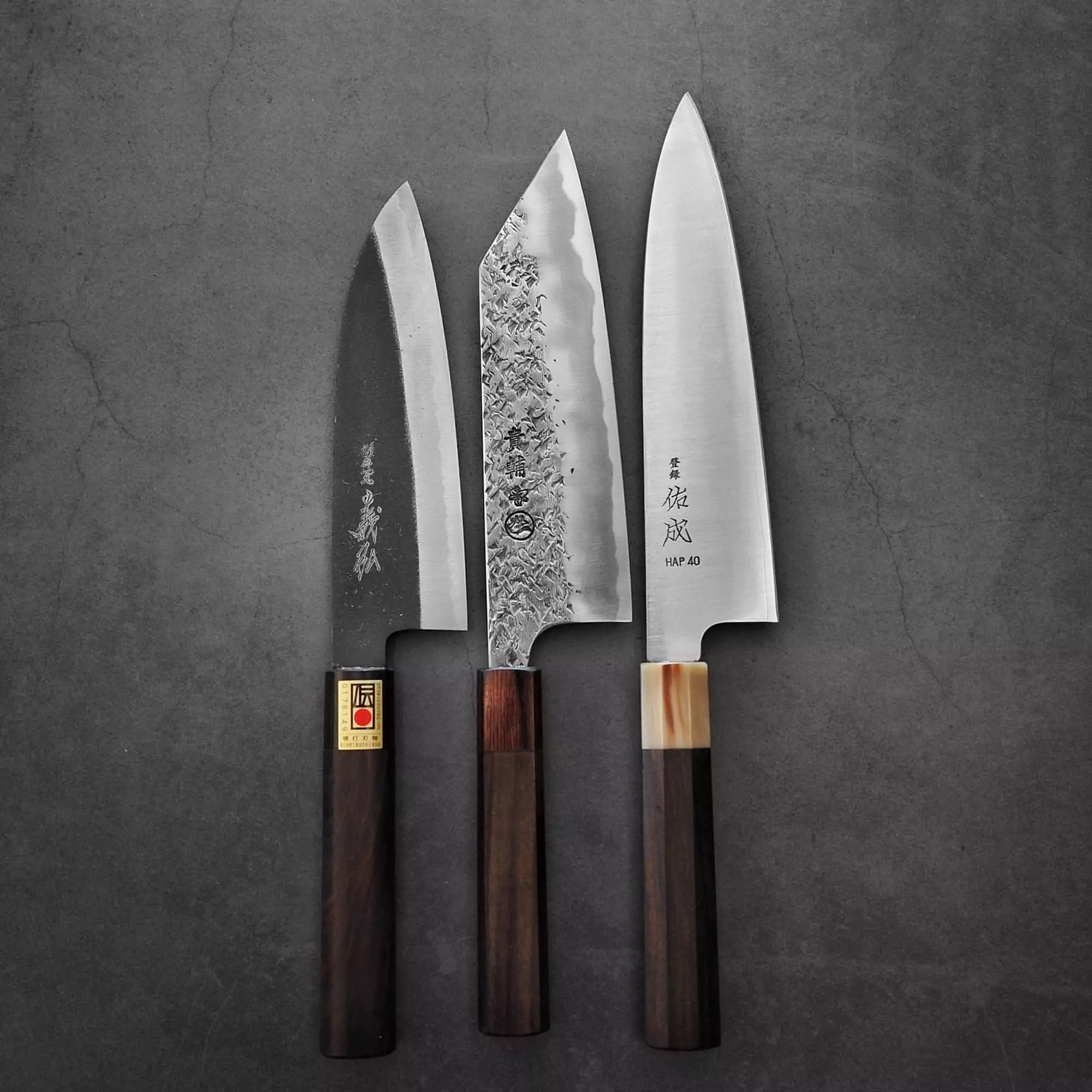 Three Japanese knives with varying blade textures and inscriptions, displayed against a grey stone background. Each knife has a wooden handle