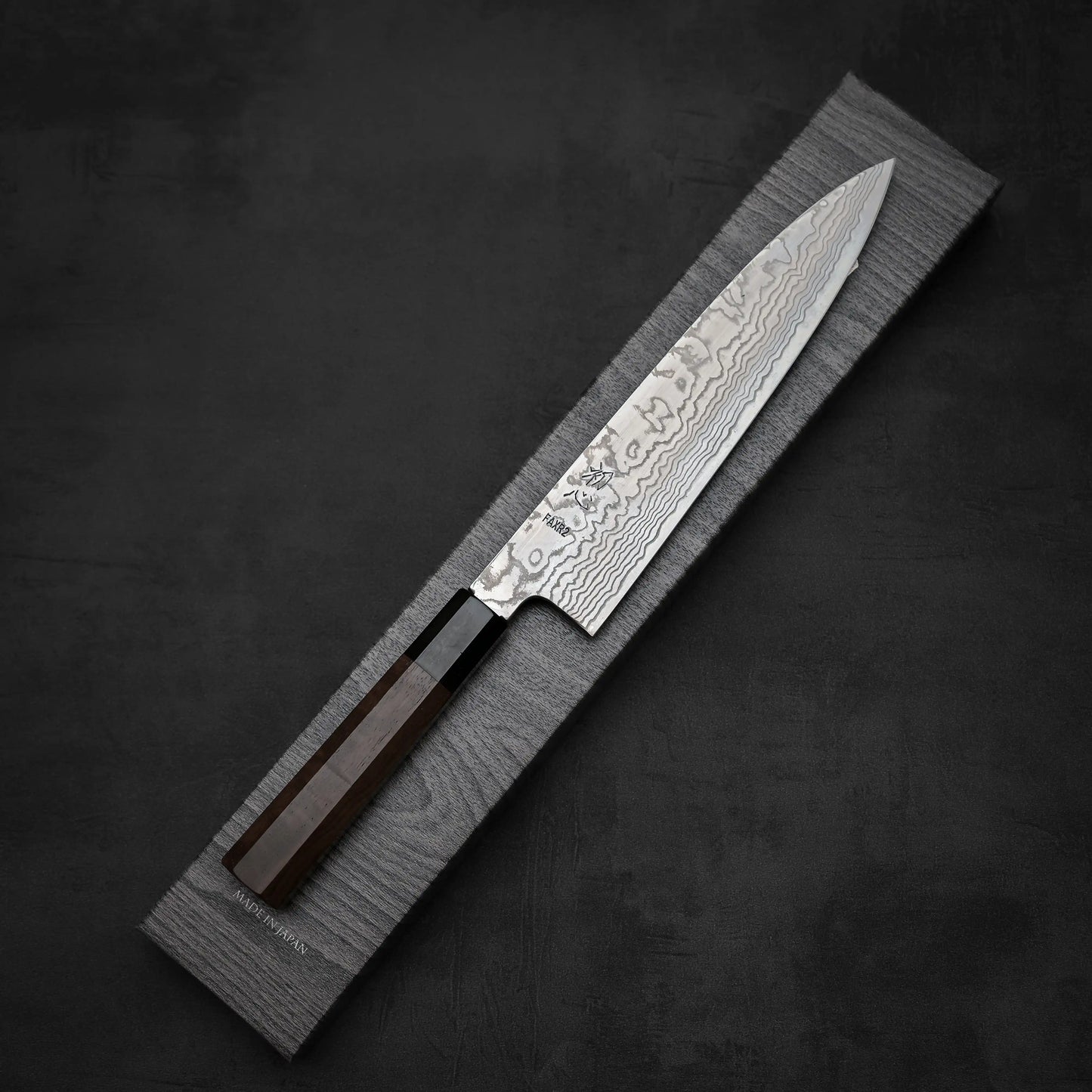 The Hatsukokoro FAXR2 Damascus gyuto 240mm, a Japanese-style chef’s knife with a patterned steel blade and dark wood handle, is displayed diagonally on a textured rectangular gray tray against a dark background.