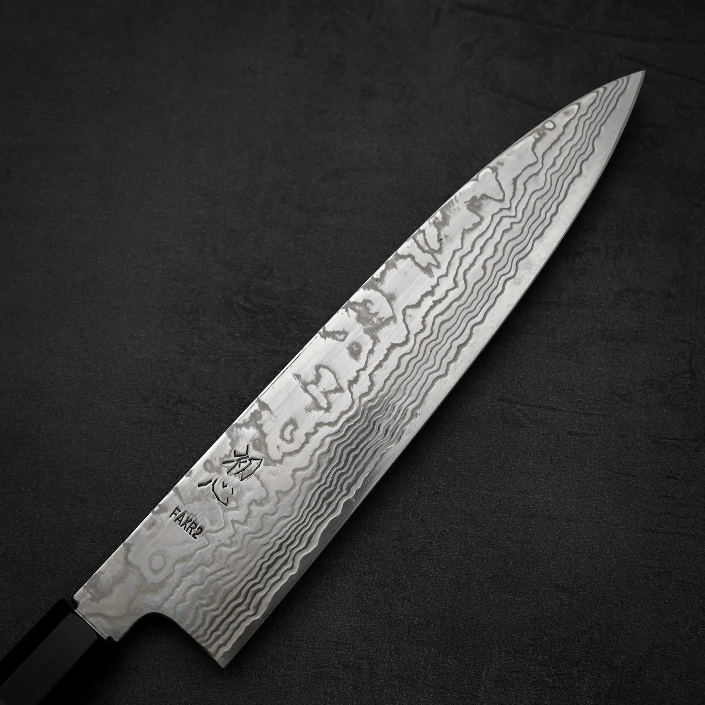 A close-up of the Hatsukokoro FAXR2 damascus gyuto 240mm chefs knife, showcasing its patterned Damascus blade and black handle, set on a dark textured surface.
