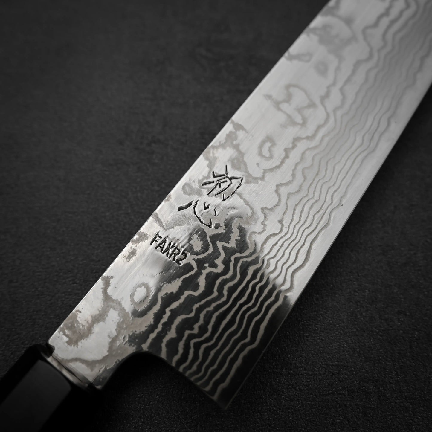 A close-up of the Hatsukokoro FAXR2 Damascus Gyuto 240mm shows its wavy-patterned blade and kanji near the handle, resting on a dark textured surface.