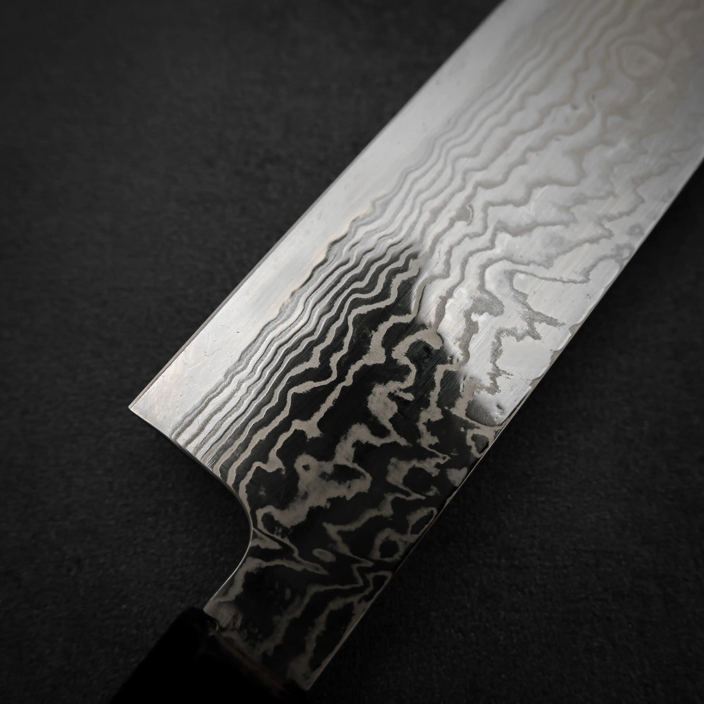Close-up of a Hatsukokoro FAXR2 damascus gyuto 240mm blade, highlighting its striking Damascus steel pattern with wavy layers and premium construction, set against a dark textured background.