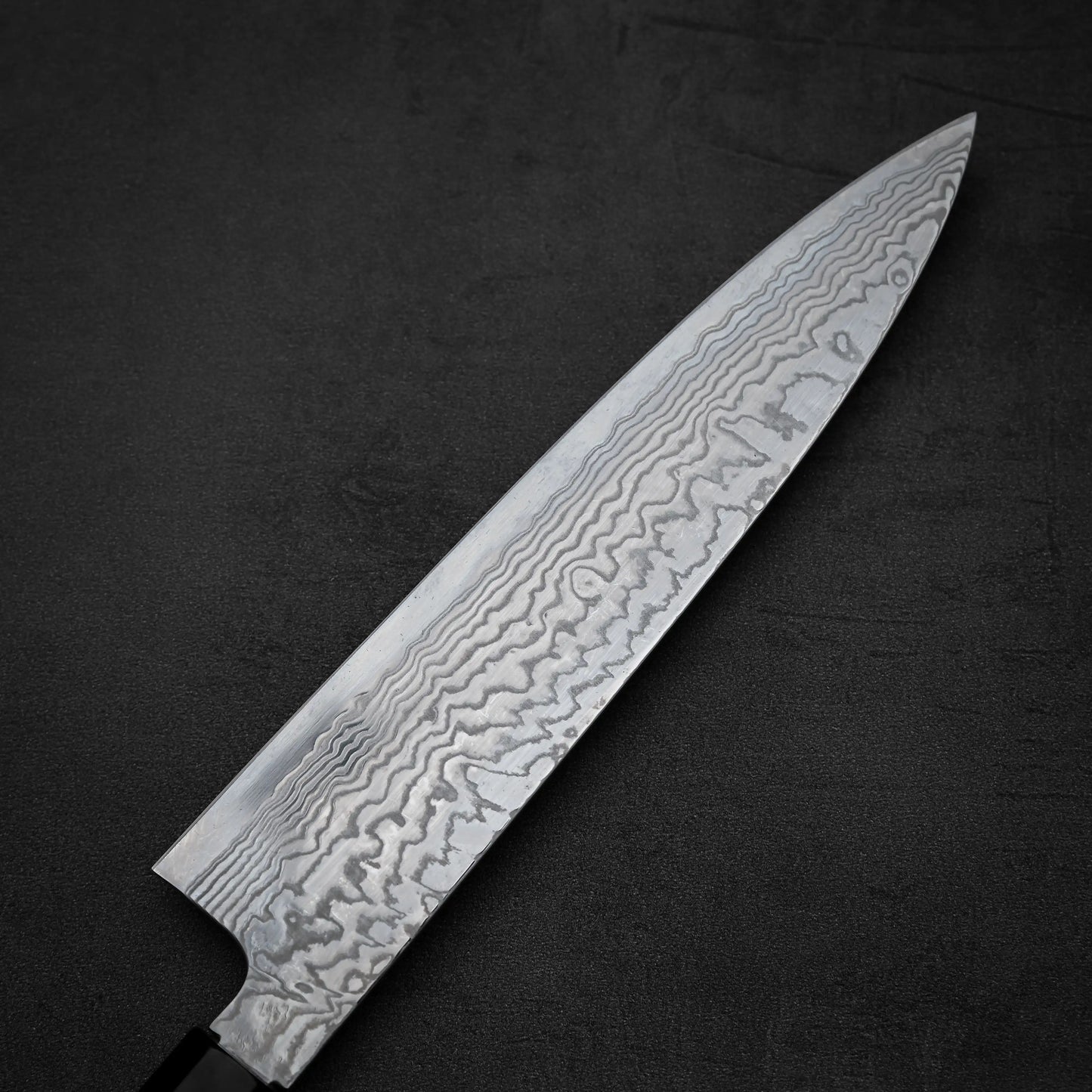 A close-up of the Hatsukokoro FAXR2 damascus gyuto 240mm knife displays its wavy, layered blade patterns on a dark, textured background.