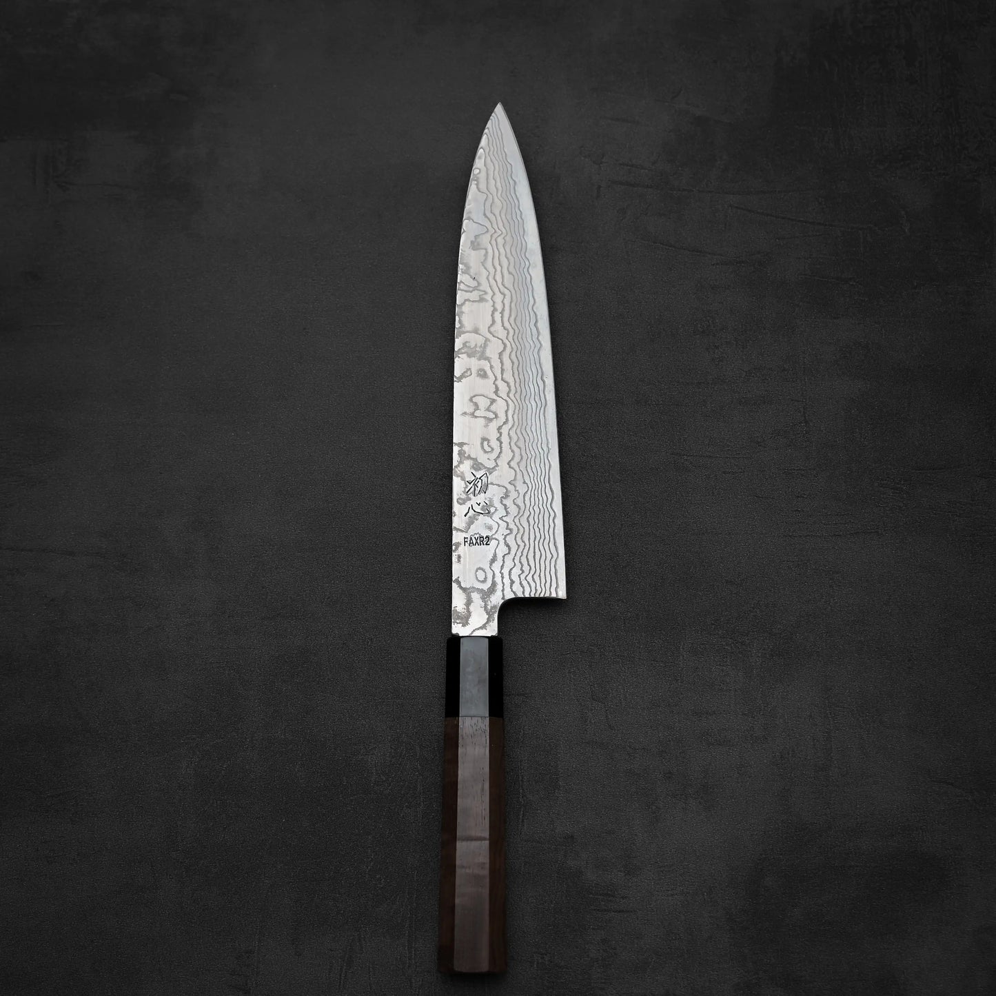 A Hatsukokoro FAXR2 Damascus Gyuto 240mm chef’s knife features a wavy, layered blade and dark wood handle, showcasing exceptional craftsmanship as it rests on a dark, textured surface.