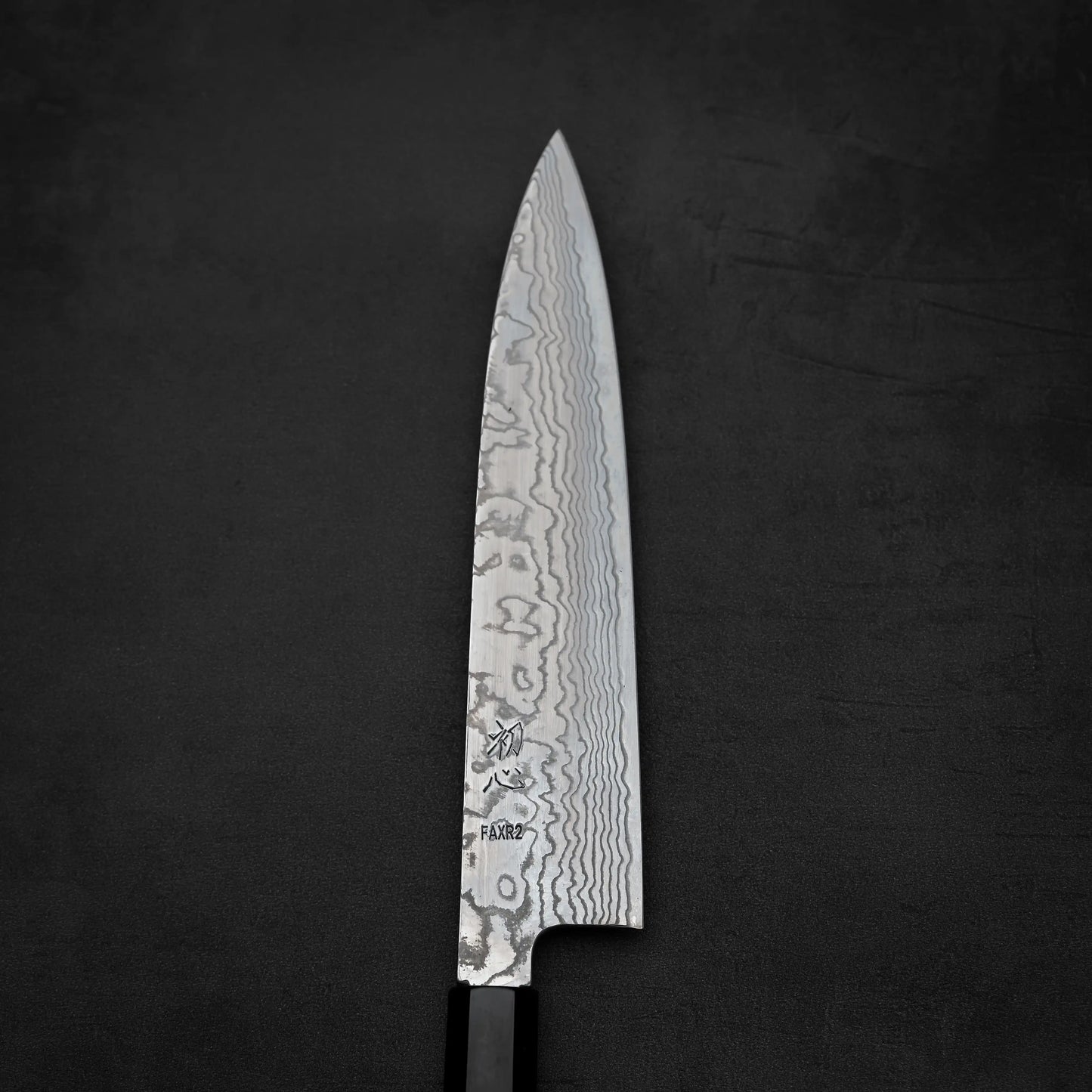 The Hatsukokoro FAXR2 damascus gyuto 240mm, featuring a patterned Damascus blade and dark handle, is shown against a black textured background.