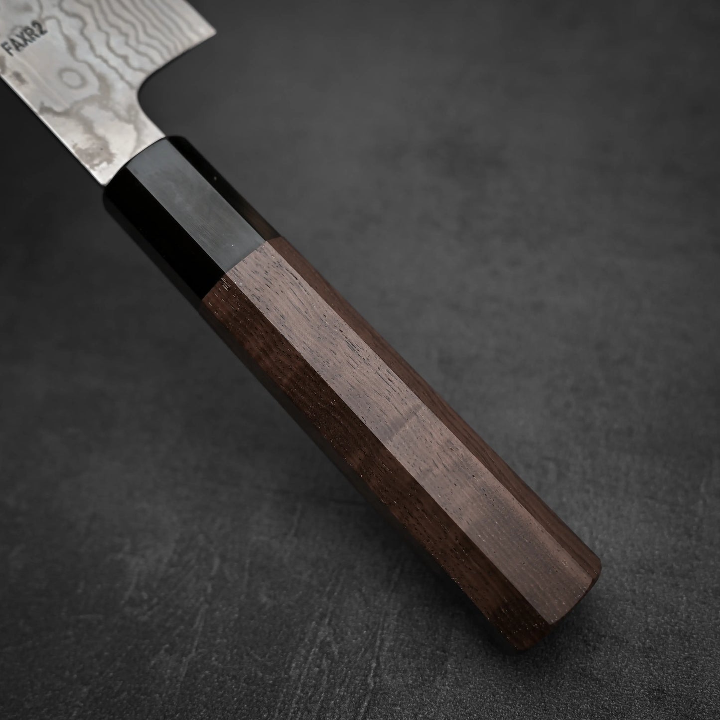 Close-up of a Hatsukokoro FAXR2 damascus gyuto 240mm handle, crafted from dark wood and black material, resting on a textured surface. The blade displays an elegant wavy pattern near the handle.