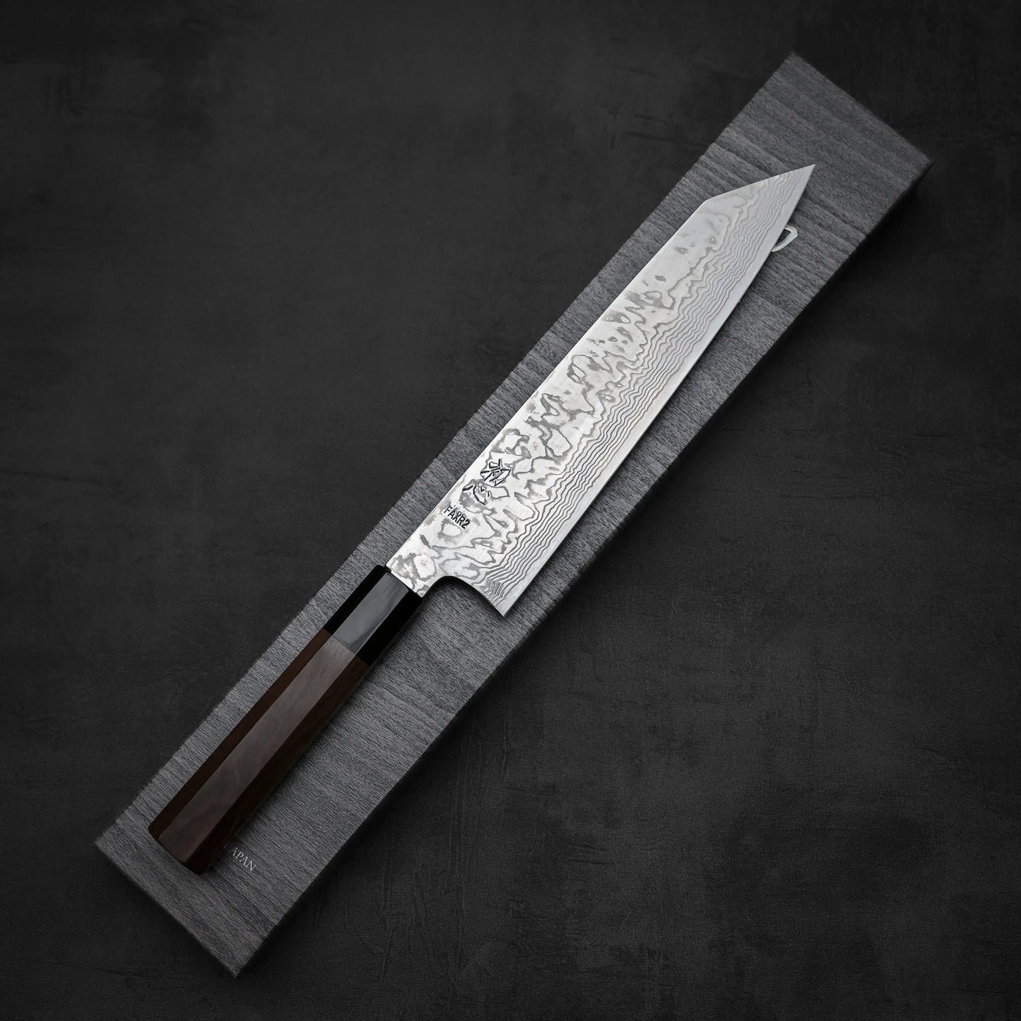 A Hatsukokoro FAXR2 damascus kiritsuke gyuto 240mm chef’s knife with a dark wooden handle lies diagonally on a rectangular gray fabric case, its blade showcasing wavy damascus patterns and engravings on a dark textured background.