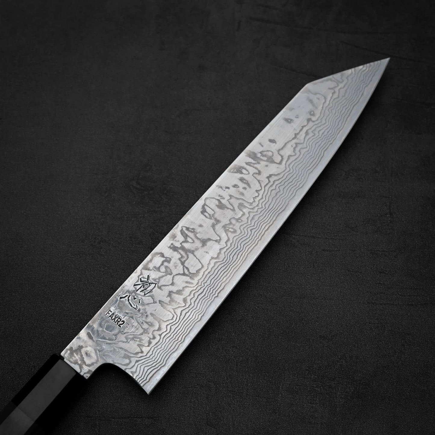 A close-up of the Hatsukokoro FAXR2 damascus kiritsuke gyuto 240mm shows its patterned Damascus steel blade with a sharp edge and etched markings, set on a dark textured surface. The knife features a black handle. Brand: Hatsukokoro.