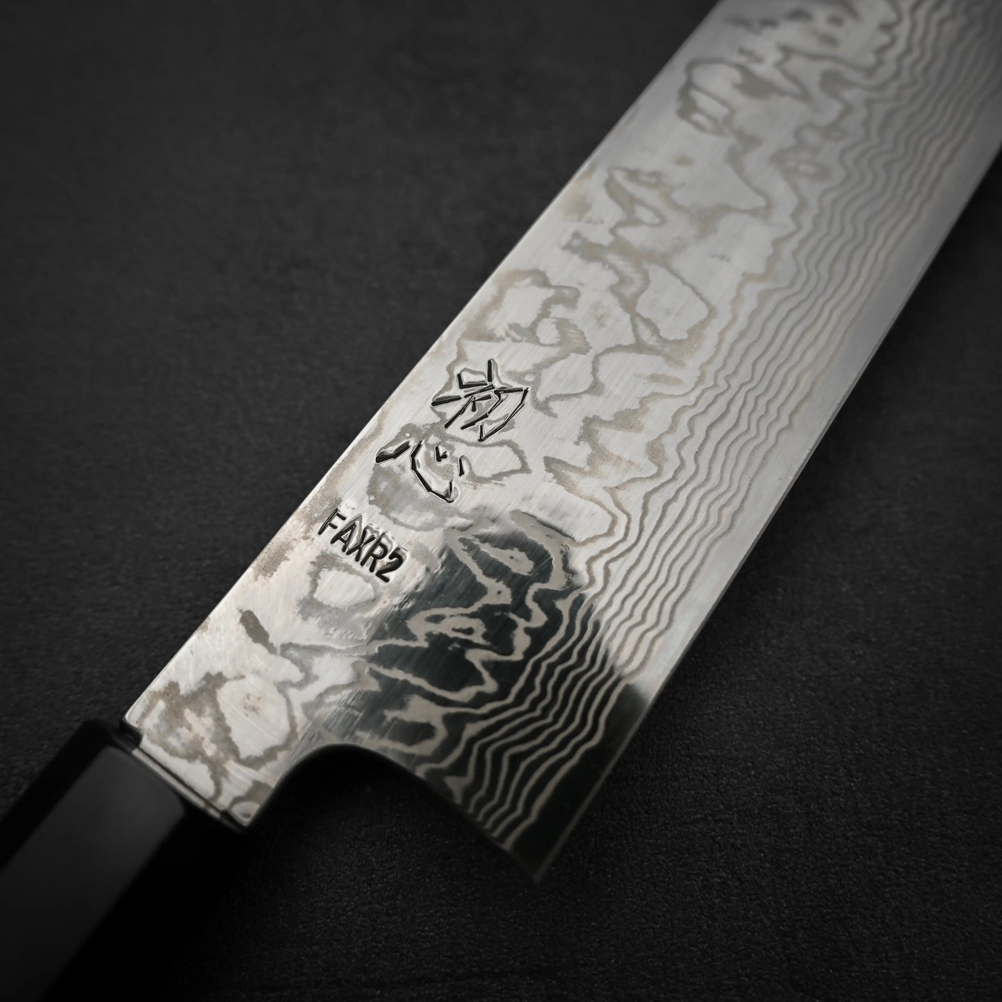 Close-up of a Hatsukokoro FAXR2 Damascus kiritsuke gyuto 240mm, showcasing its detailed blade pattern, Japanese characters, and the brand name stamped near the handle on a dark surface.