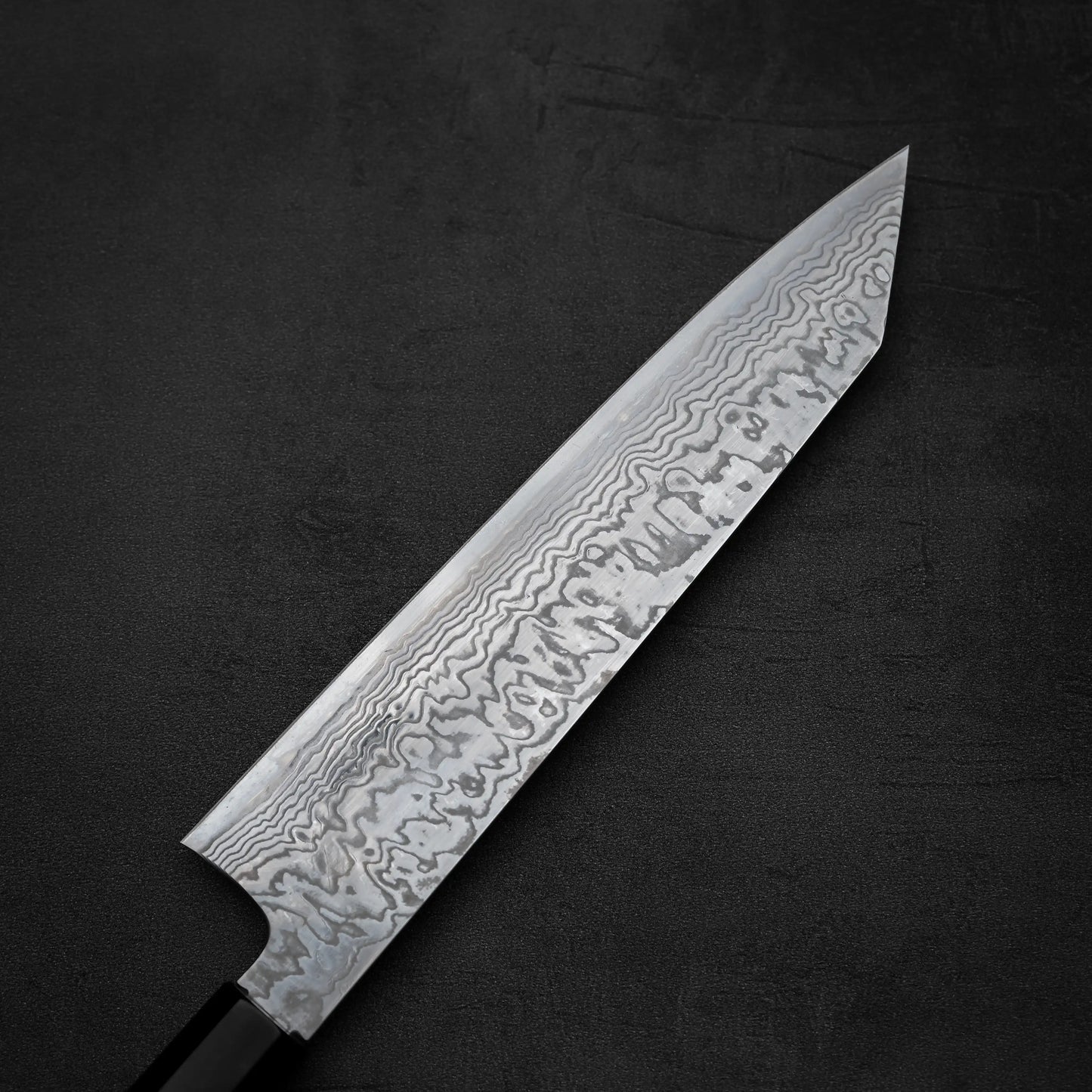 A close-up of the Hatsukokoro FAXR2 damascus kiritsuke gyuto 240mm knife with a patterned blade and black handle, displayed on a dark textured surface.
