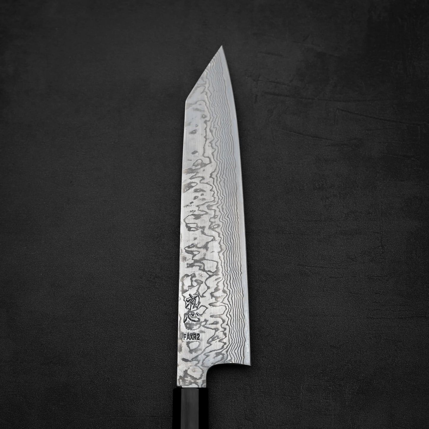 The Hatsukokoro FAXR2 damascus kiritsuke gyuto 240mm, a Japanese-style chef knife by Hatsukokoro with a patterned Damascus blade and black handle, rests on a dark textured surface.