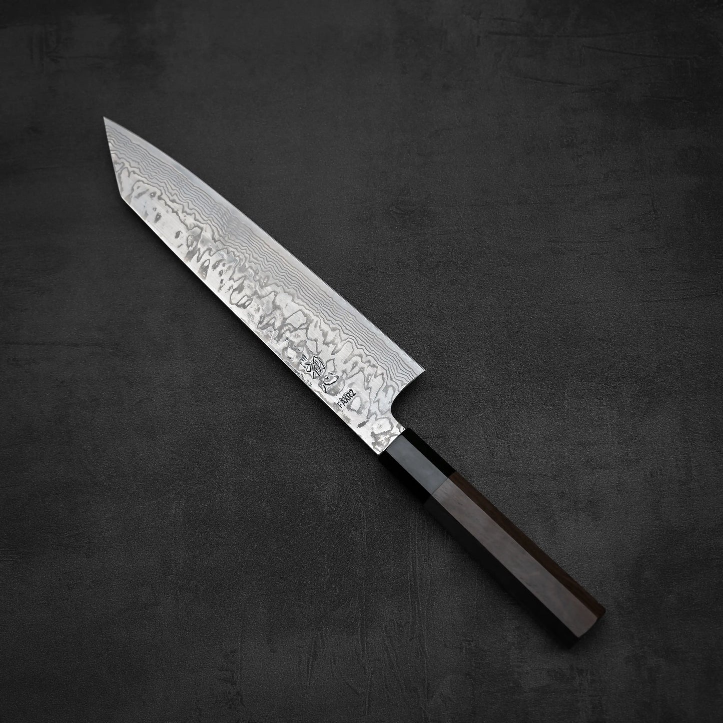 A Hatsukokoro FAXR2 damascus kiritsuke gyuto 240mm chef’s knife with a dark wooden handle rests on a dark textured surface.
