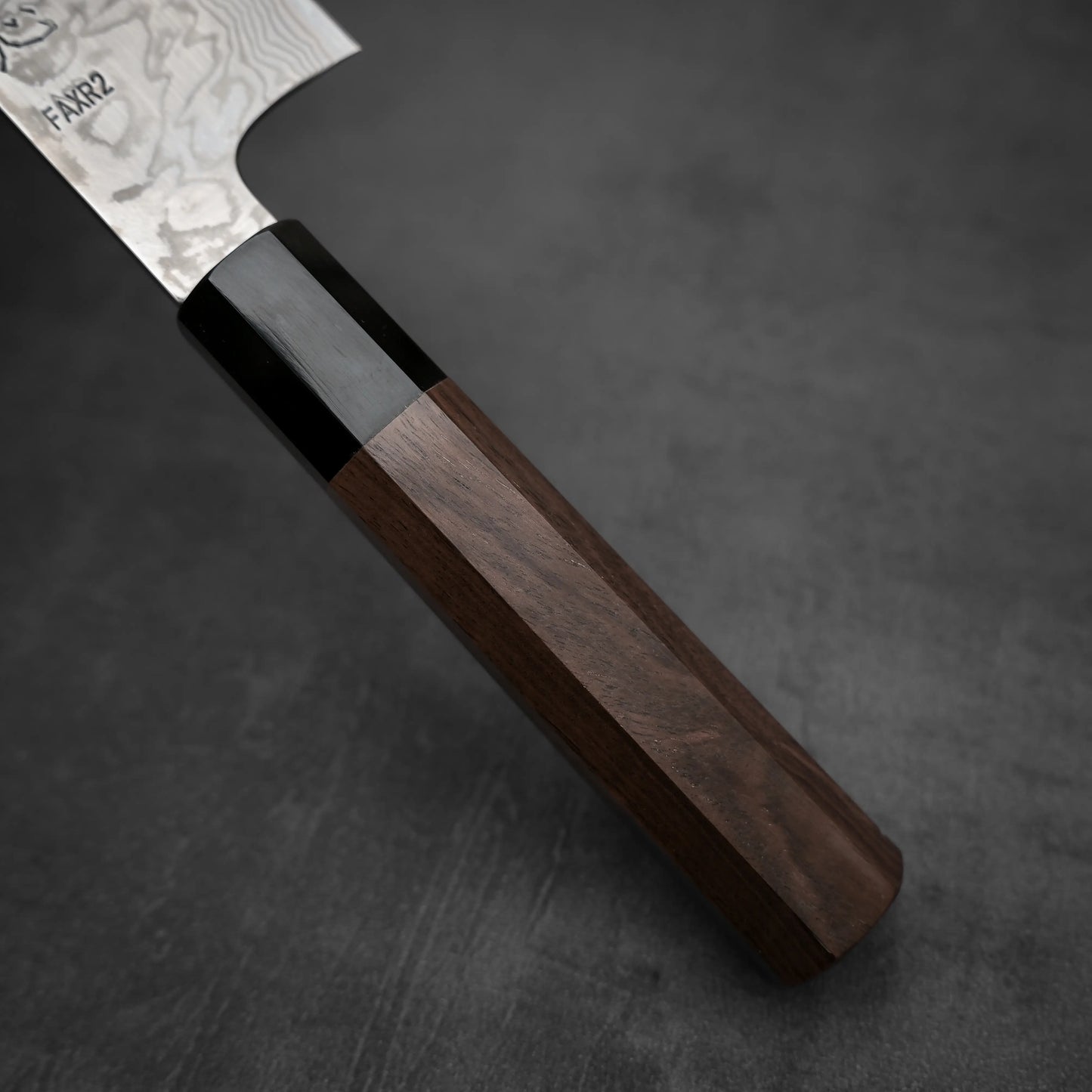 Close-up of a Hatsukokoro FAXR2 damascus kiritsuke gyuto 240mm handle made of dark wood with a glossy black bolster, resting on a textured surface. The patterned blade is partially visible in the upper left corner.