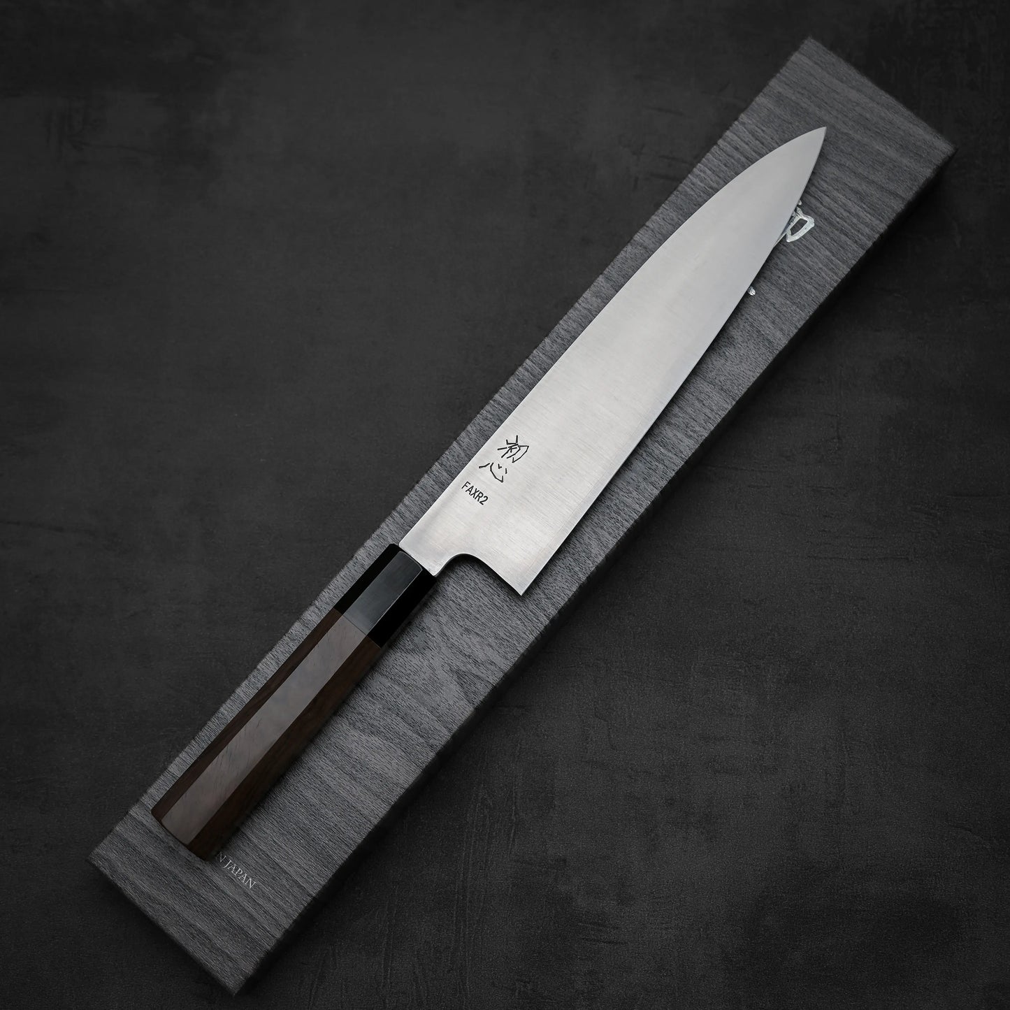 The Hatsukokoro FAXR2 gyuto 240mm, a Japanese knife with a wooden handle and polished SG2 stainless steel blade known for excellent edge retention, is shown on a rectangular gray textured box against a dark background.