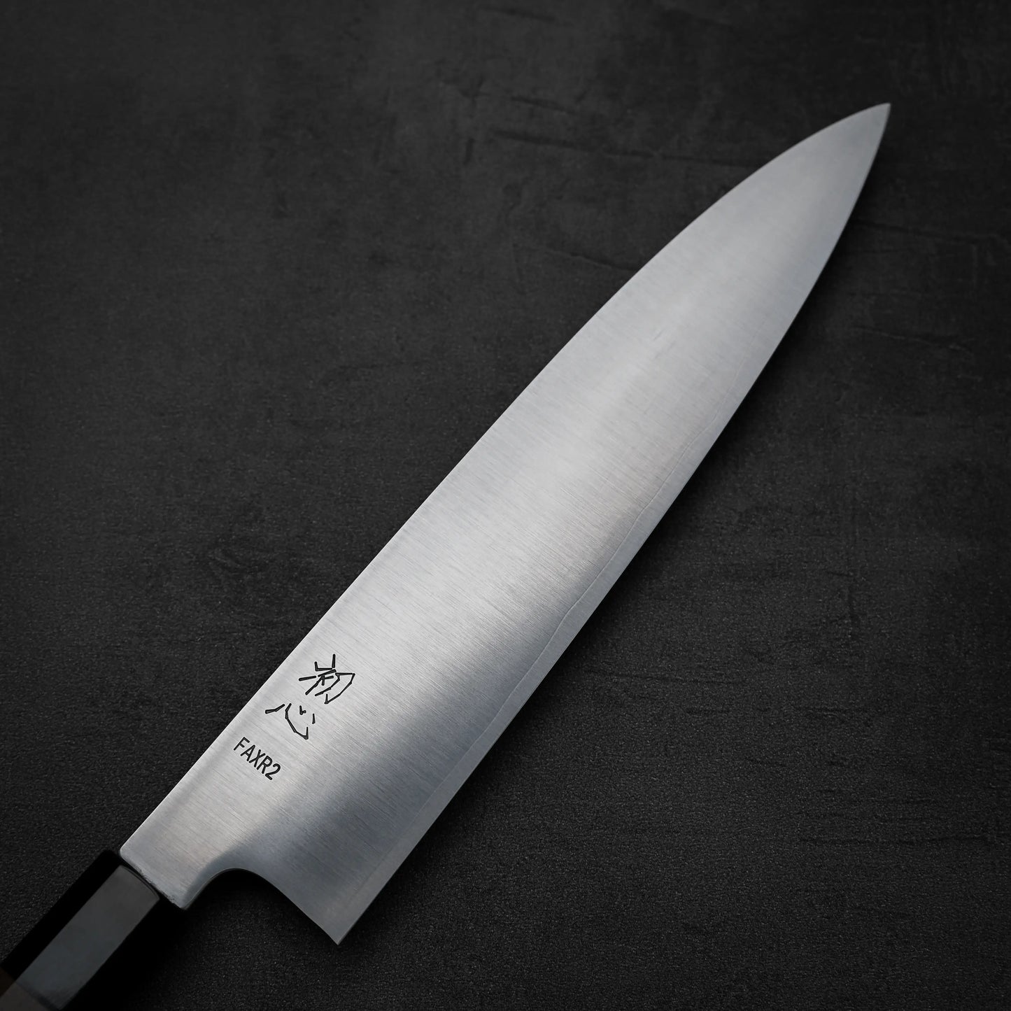 A close-up of the Hatsukokoro FAXR2 gyuto 240mm knife, featuring Japanese characters and FAXR2 engraved on its blade, by Hatsukokoro, displayed on a black textured background.