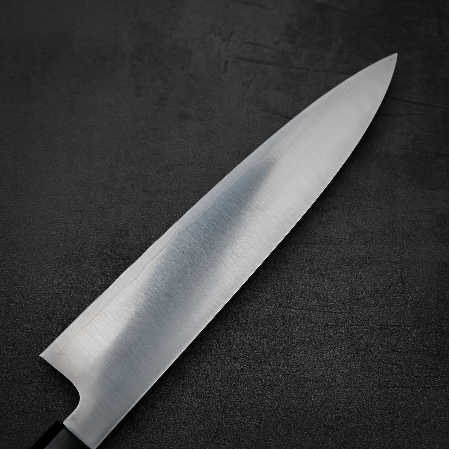 A close-up of the Hatsukokoro FAXR2 gyuto 240mm by Hatsukokoro, featuring a sharp, reflective stainless steel blade and black handle, displayed on a dark textured surface.