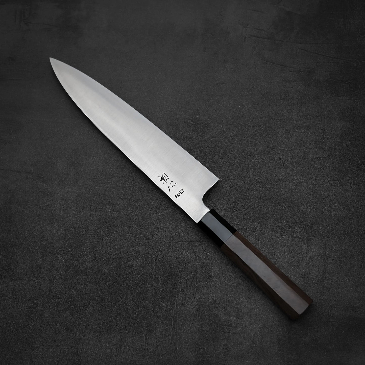 A Hatsukokoro FAXR2 gyuto 240mm knife with a lustrous stainless steel blade and dark wood handle lies on textured gray. Japanese characters and Hatsukokoro branding are engraved on the blade, reflecting its outstanding edge retention.