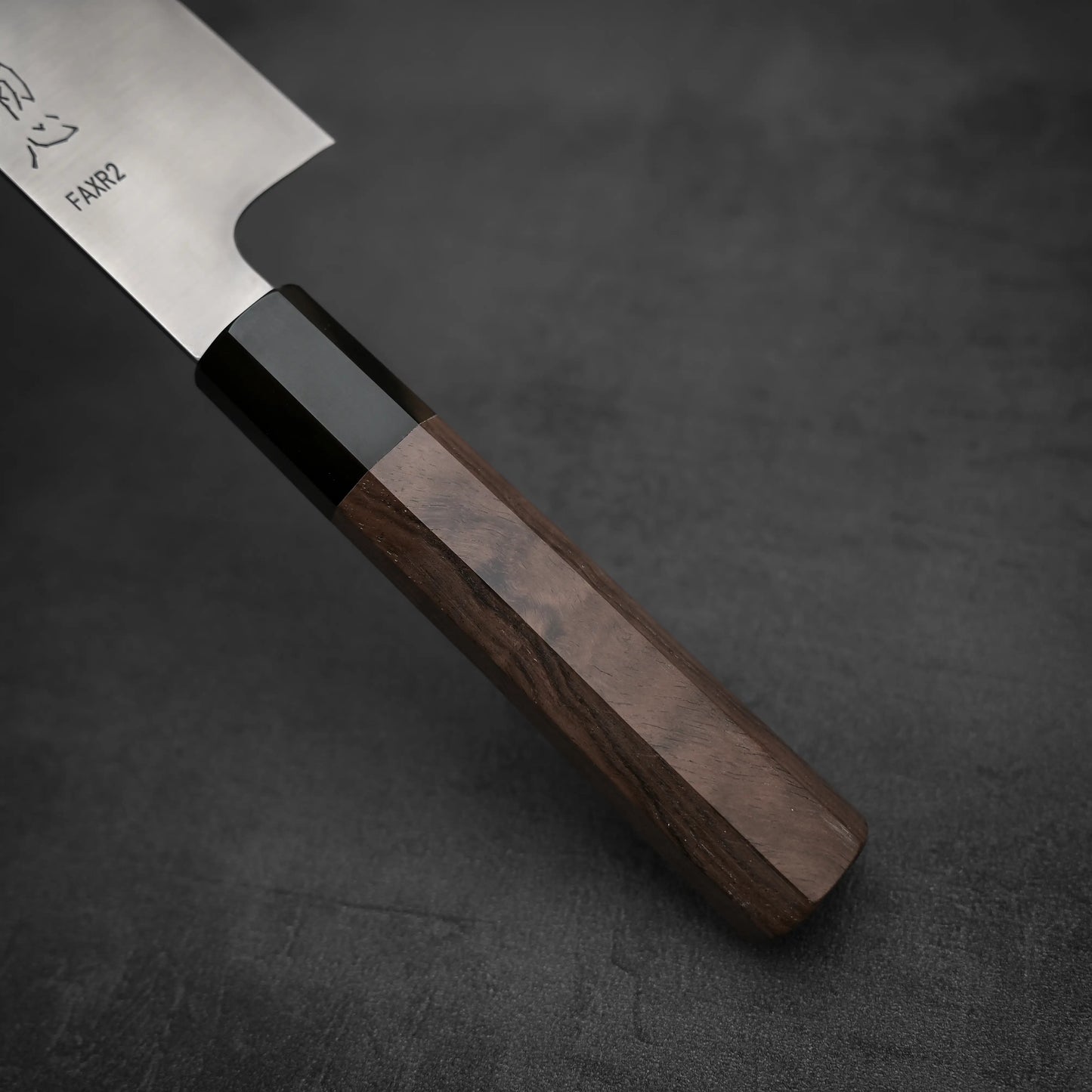 Close-up of the Hatsukokoro FAXR2 gyuto 240mm by Hatsukokoro, showcasing its polished dark wooden handle and SG2 stainless steel blade, renowned for excellent edge retention, set against a dark gray textured background.