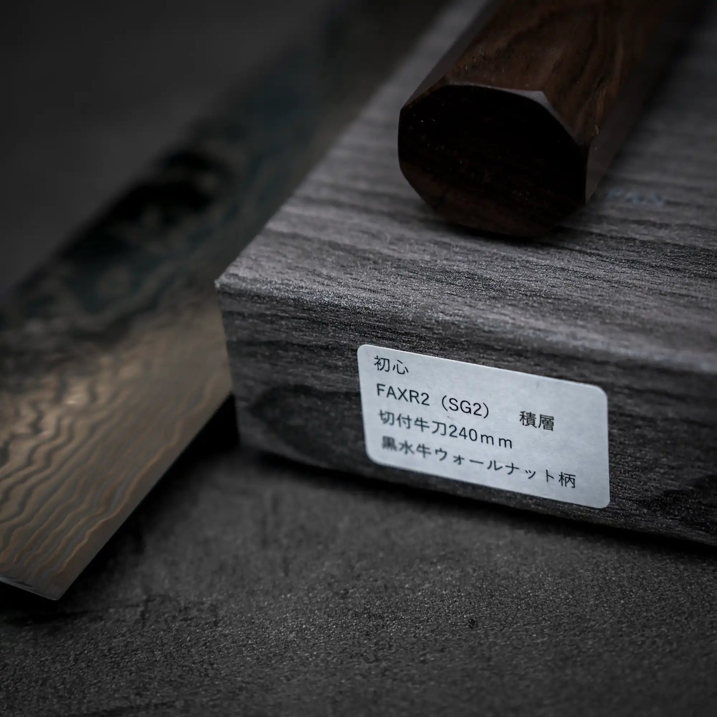 A close-up of a Hatsukokoro FAXR2 damascus gyuto 240mm Japanese kitchen knife with a patterned blade and wooden handle, resting on a gray wooden box labeled with Japanese text and product details.