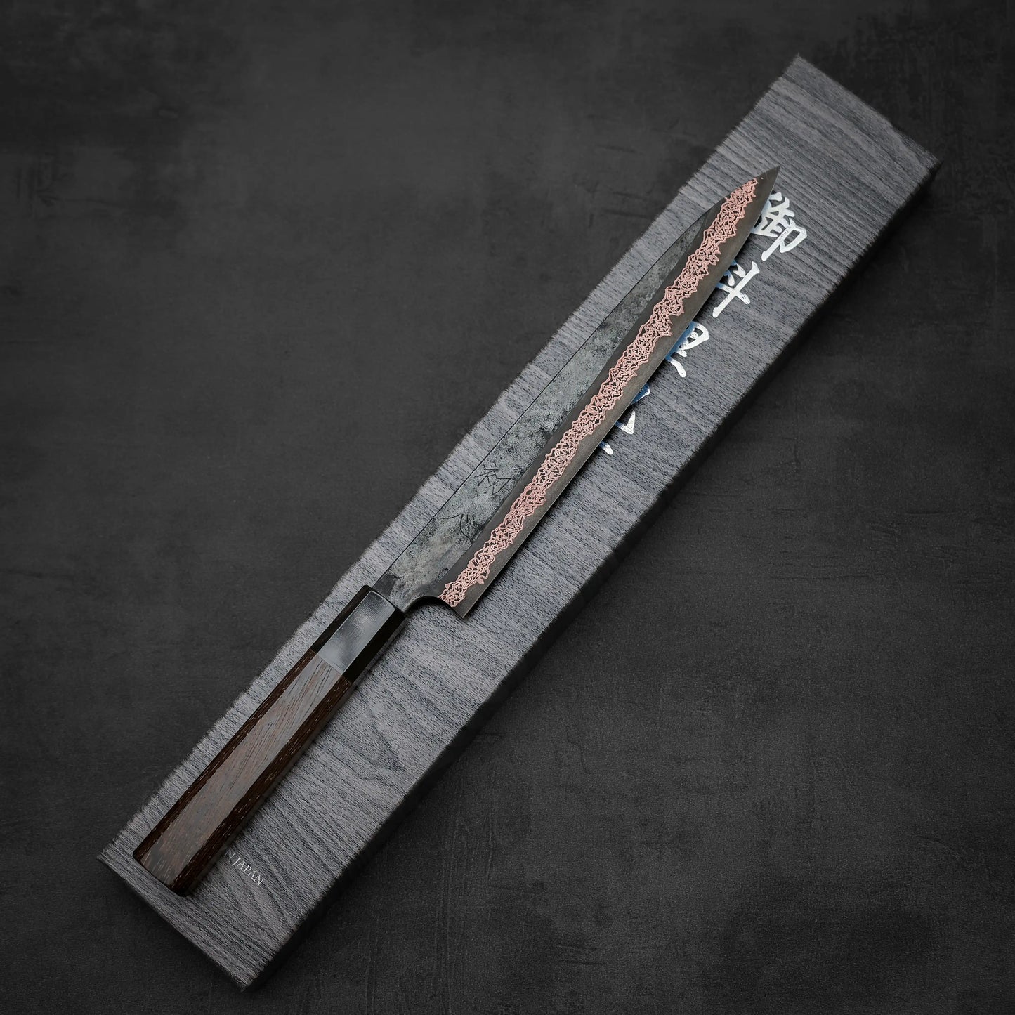 The Hatsukokoro Yorokobi SLD kurouchi copper damascus sujihiki 240mm, featuring a dark wooden handle and damascus-patterned blade, rests diagonally on a gray box with white Japanese characters against a dark, textured background.