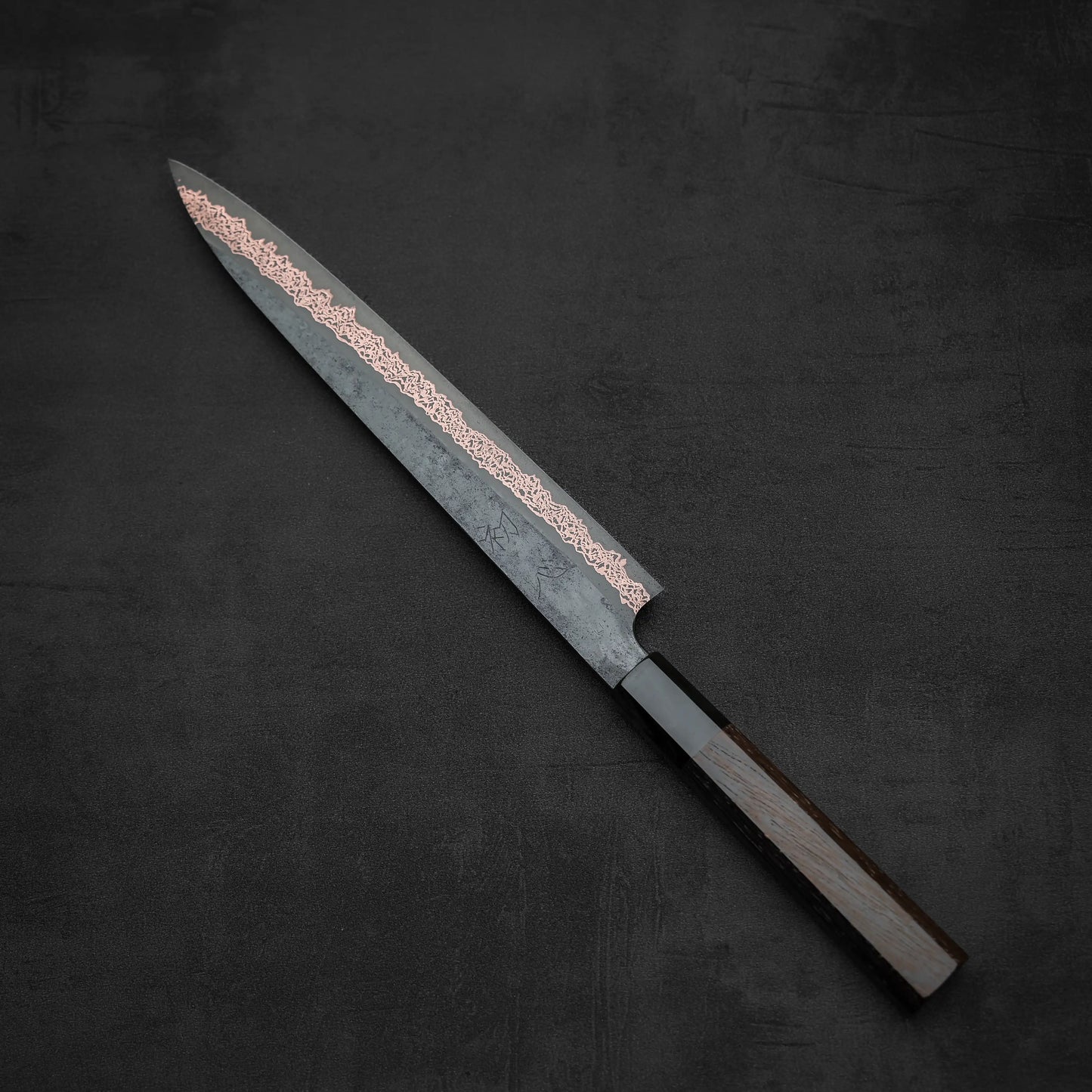 The Hatsukokoro Yorokobi SLD kurouchi copper damascus sujihiki 240mm by Hatsukokoro features a dark wooden handle and a long, pointed patterned blade, shown on a dark textured surface.