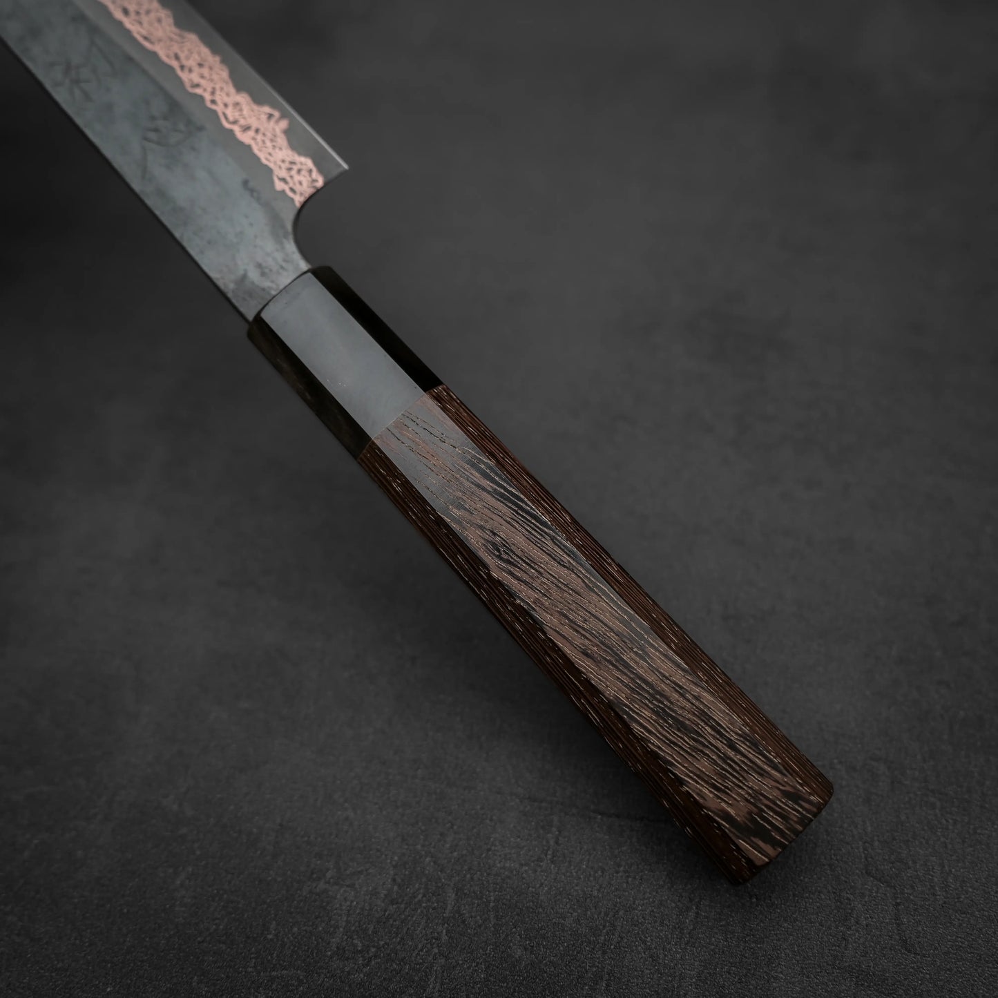 Close-up of the Hatsukokoro Yorokobi SLD kurouchi copper damascus sujihiki 240mm by Hatsukokoro, featuring a dark wooden handle and patterned blade, resting on a dark textured surface.