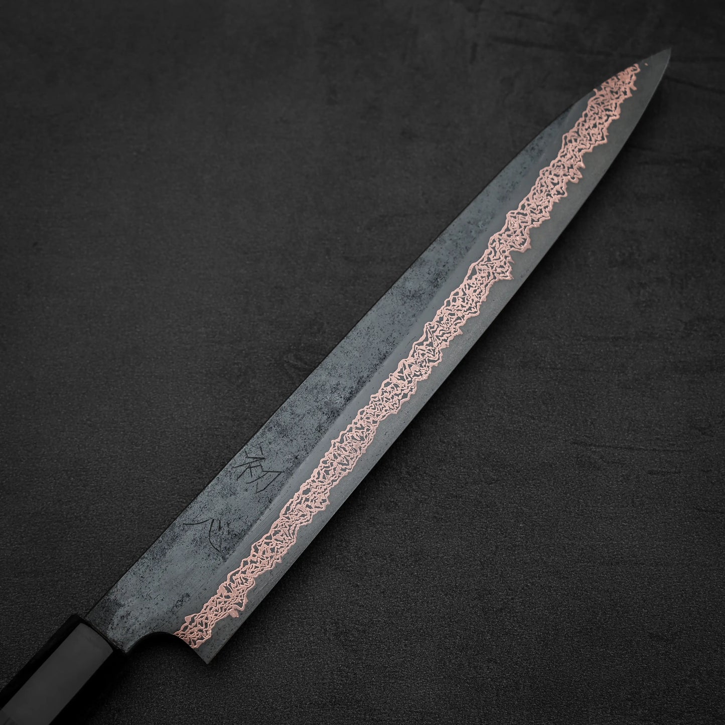 The Hatsukokoro Yorokobi SLD kurouchi copper damascus sujihiki 240mm features a sleek Japanese blade with a dark kurouchi finish, copper damascus pattern near the edge, and a black handle—showcasing Hatsukokoro’s elegant craftsmanship.