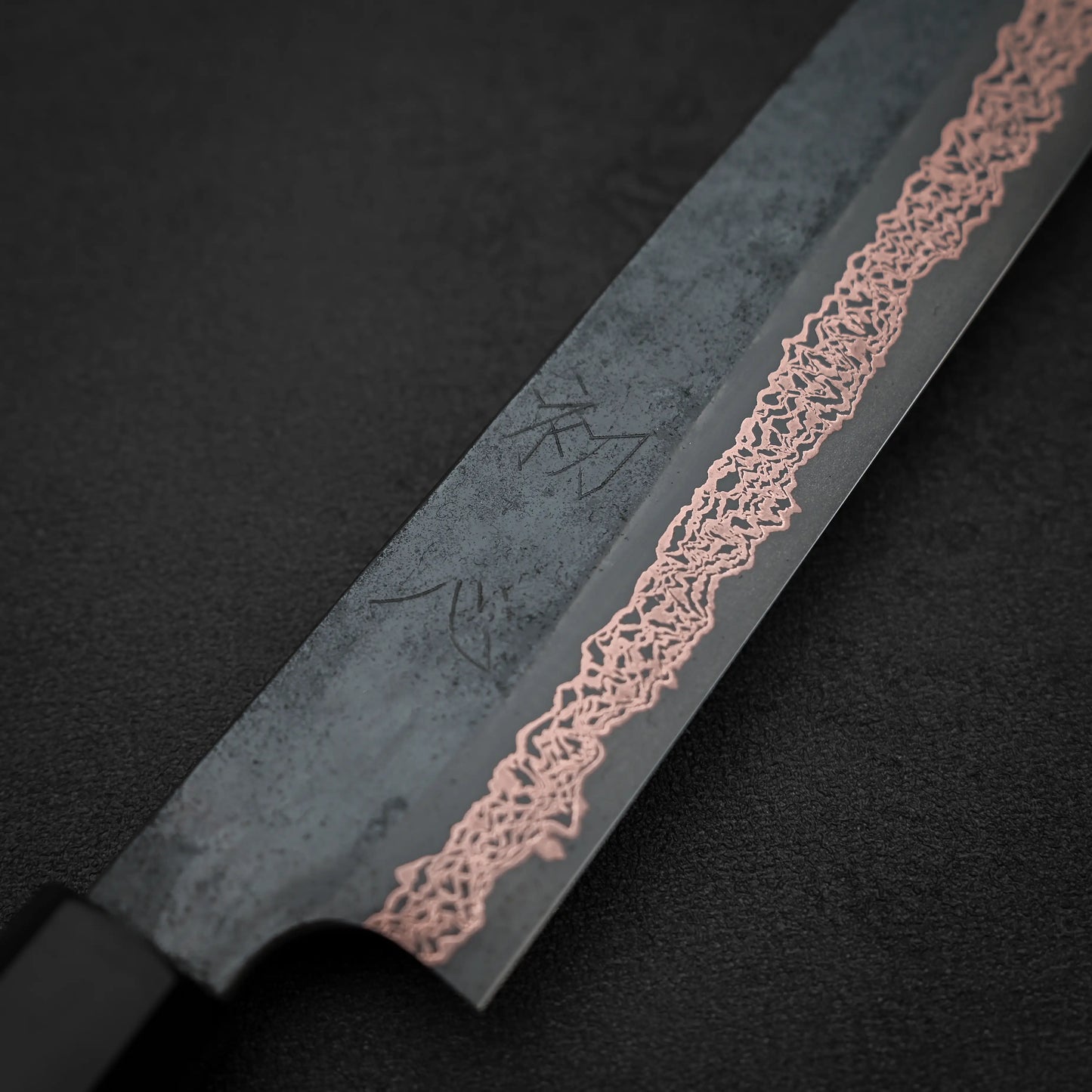 A close-up of the Hatsukokoro Yorokobi SLD kurouchi copper damascus sujihiki 240mm by Hatsukokoro, featuring its textured black damascus blade with a wavy copper pattern near the edge, set against a dark background.