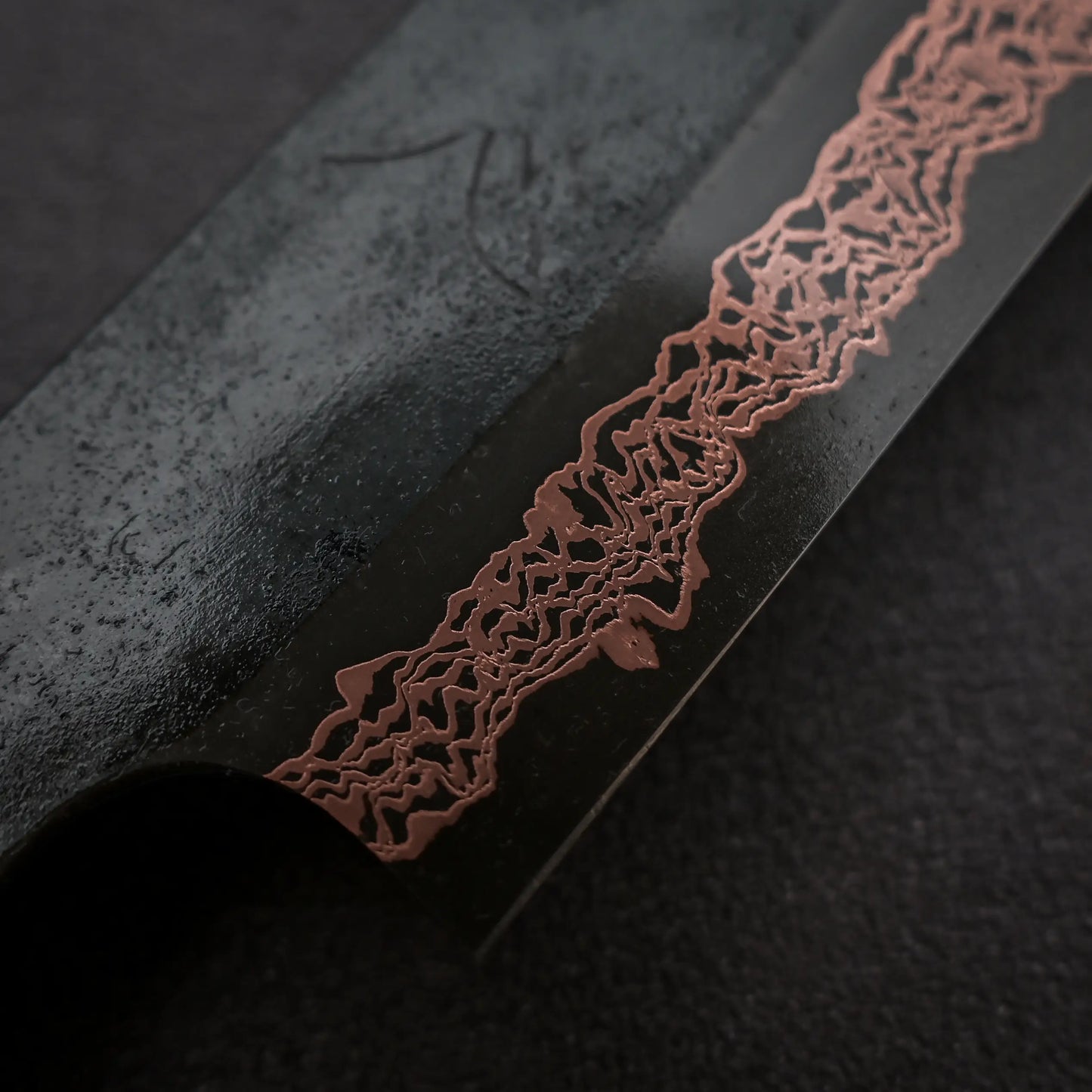 Close-up of the Hatsukokoro Yorokobi SLD kurouchi copper damascus sujihiki 240mm by Hatsukokoro, showing its dark blade with a copper-colored, wavy damascus pattern along the edge, resting on a textured black surface.