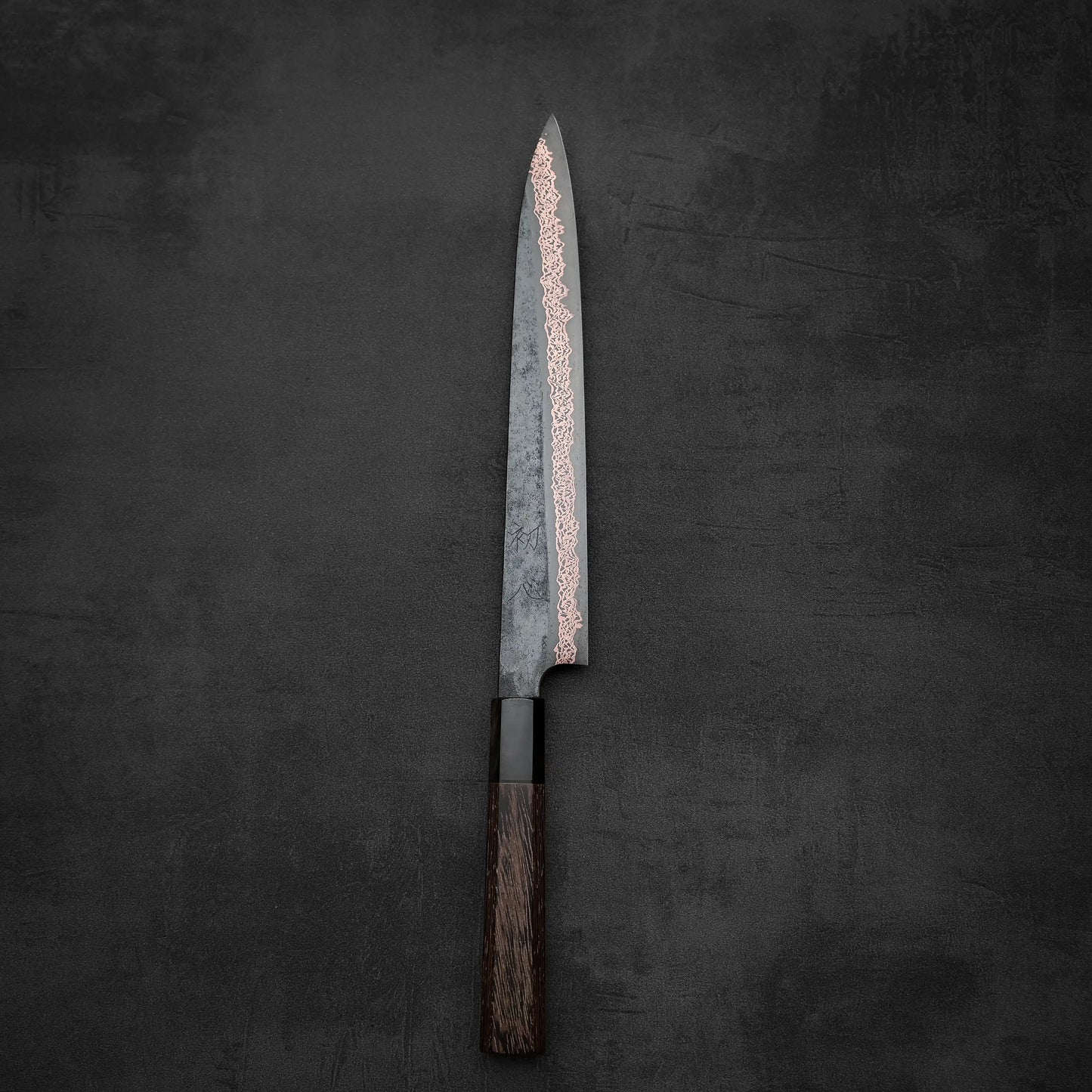 The Hatsukokoro Yorokobi SLD kurouchi copper damascus sujihiki 240mm by Hatsukokoro, featuring a dark wooden handle and damascus-patterned blade, stands vertically on a dark, textured surface.