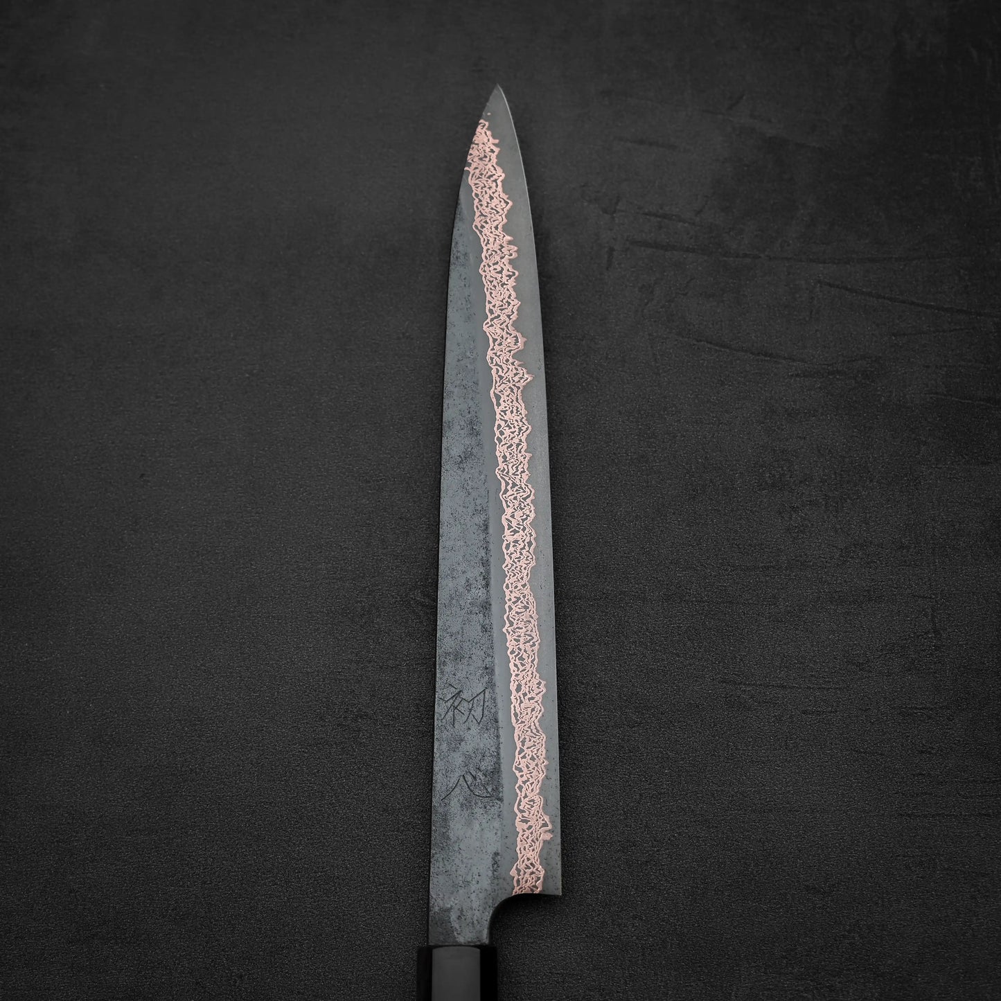The Hatsukokoro Yorokobi SLD kurouchi copper damascus sujihiki 240mm by Hatsukokoro, with a wavy damascus pattern on its blade, is displayed against a dark textured background.