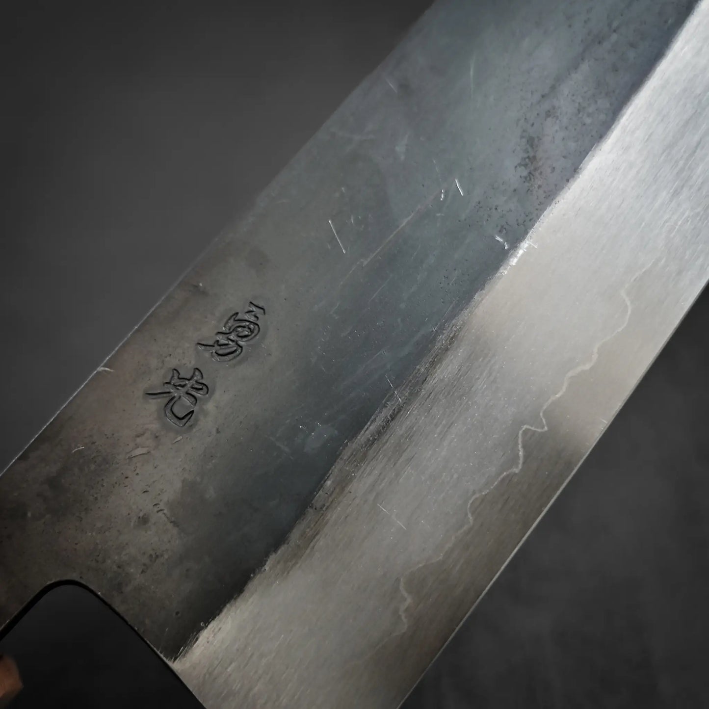 Close-up of the Isamitsu kurouchi shirogami#1 gyuto 210mm knife blade by Isamitsu, showing Japanese etching, a subtle brushed metal pattern, signs of use, and a sharp edged finish.