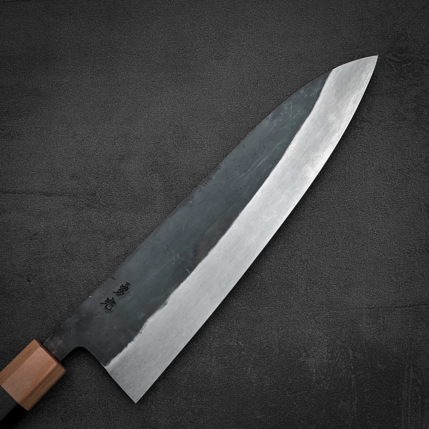 The Isamitsu kurouchi shirogami#1 gyuto 210mm knife by Isamitsu, with a dark kurouchi blade, wooden handle, and Japanese characters near the handle, rests on a textured dark surface.