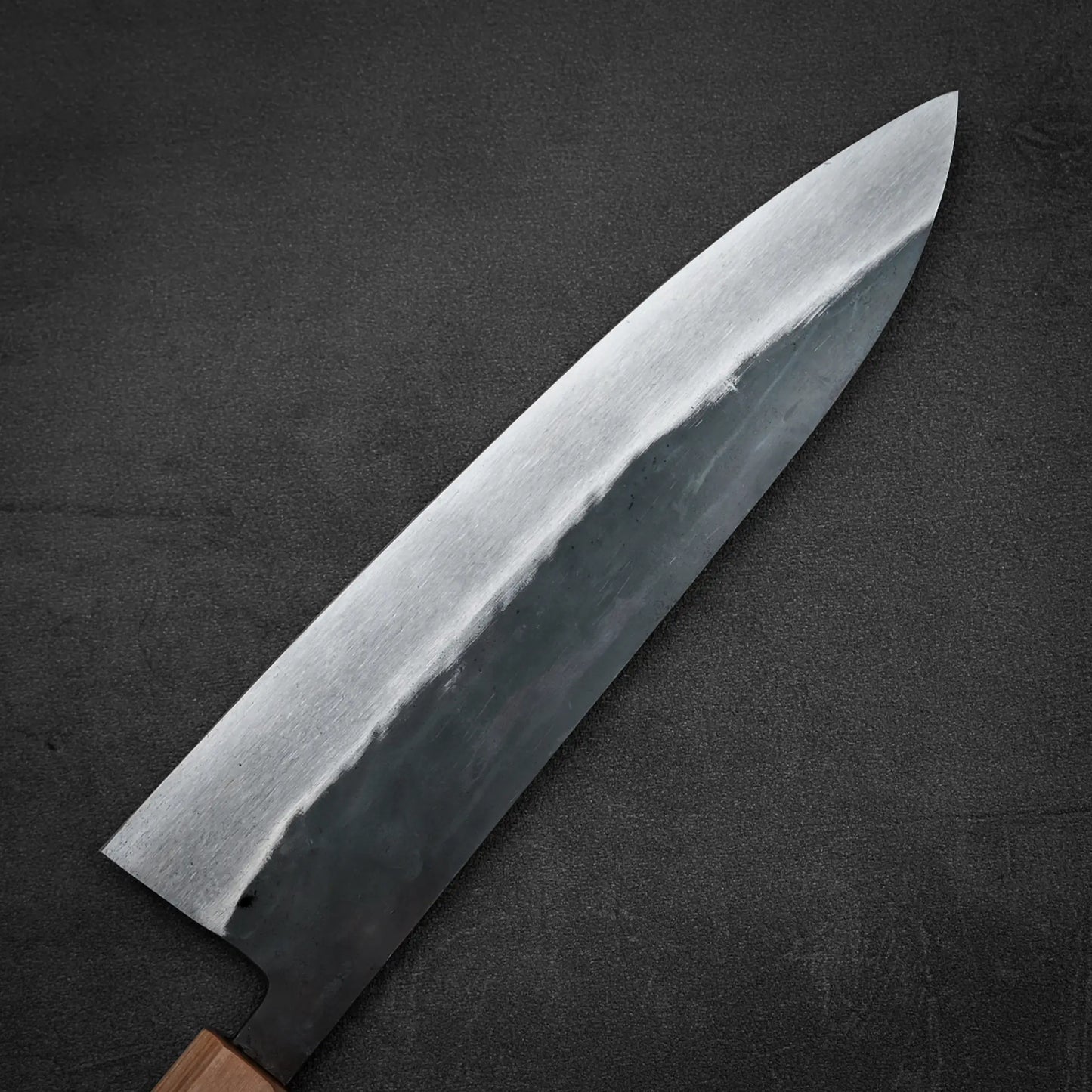 A close-up of the Isamitsu kurouchi shirogami#1 gyuto 210mm Japanese knife by Isamitsu, featuring a polished steel blade and dark matte handle, displayed on a textured black surface.